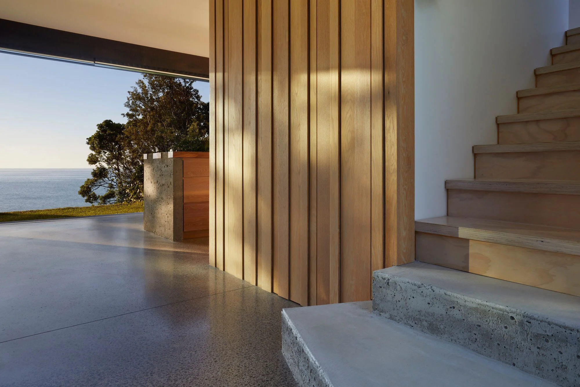 Modern interior with wooden wall paneling, concrete and wooden staircase, and large open window overlooking the ocean and trees at sunset.