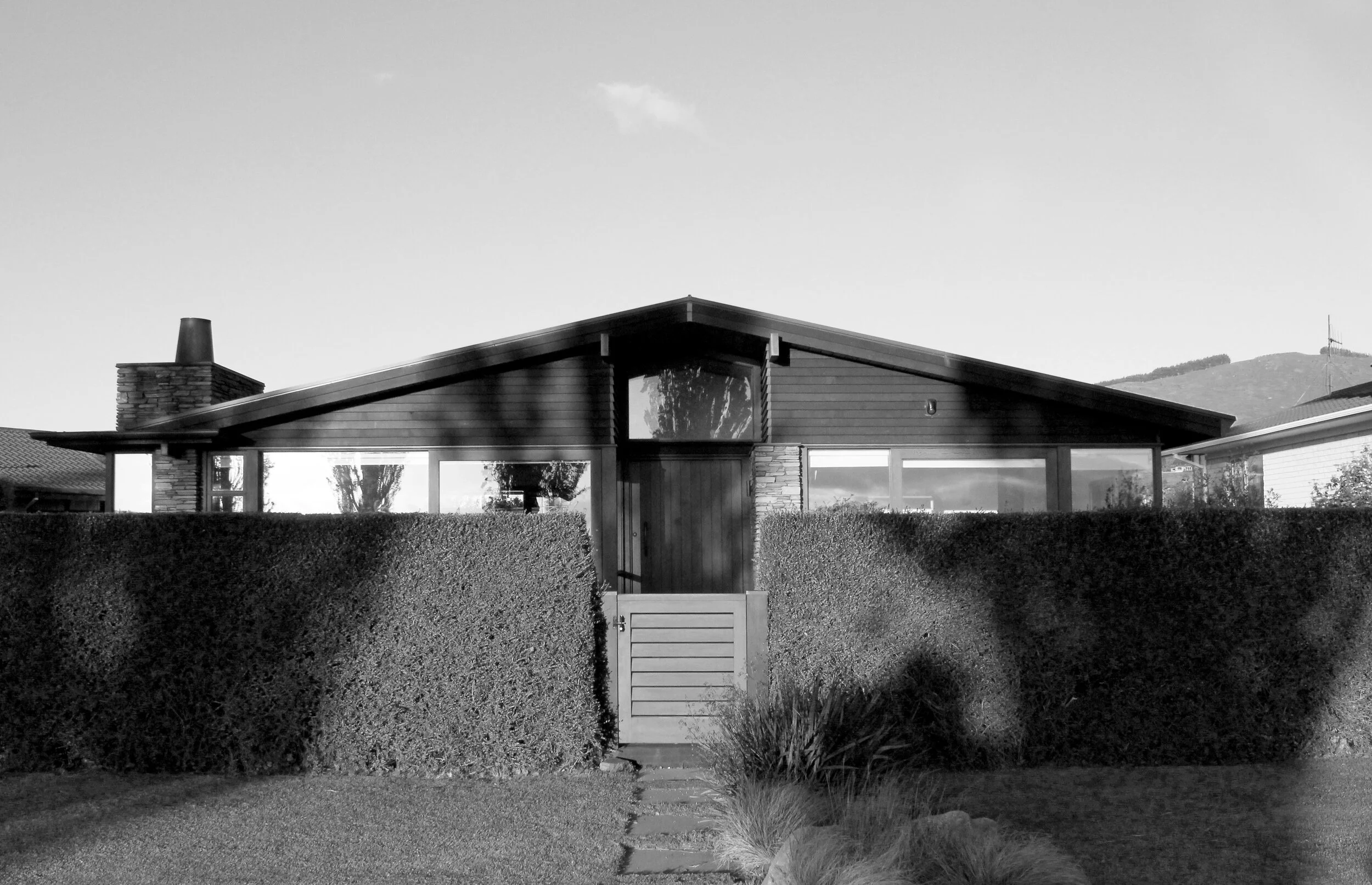 Black and white photo of a modern house with a sloped roof, large windows, a chimney, and surrounded by a tall hedge and small gate.