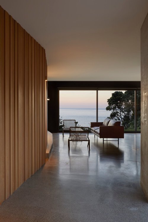 Living room with large windows overlooking a body of water and trees, furnished with a sofa and chairs, with a view of the sunset.