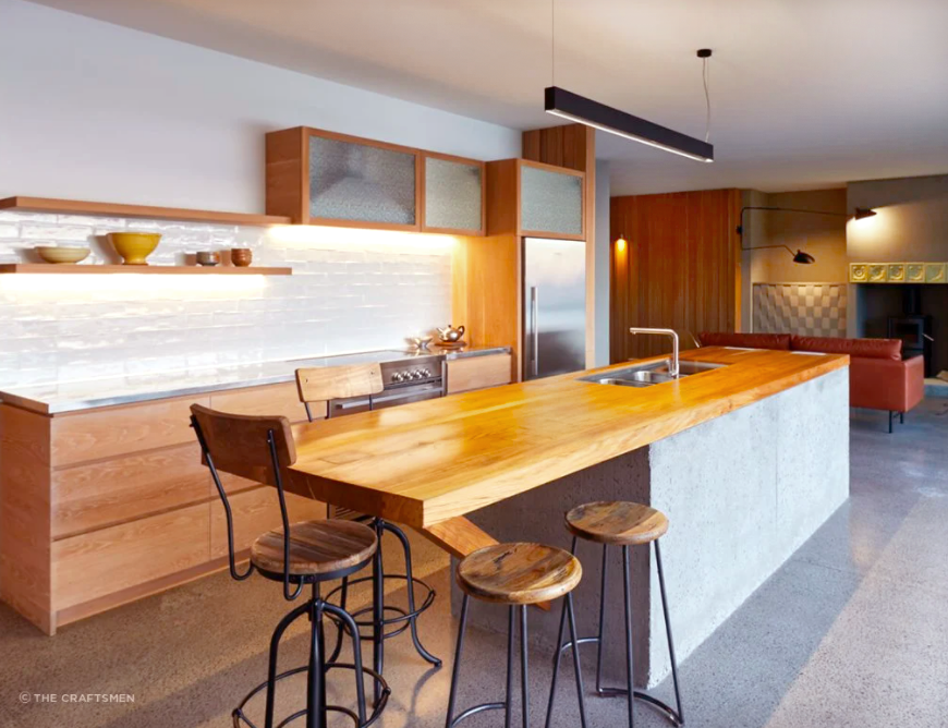 Modern kitchen with wooden island countertop, three bar stools, white brick backsplash, open shelving, stainless steel appliances, and a living area with a brown couch in the background.