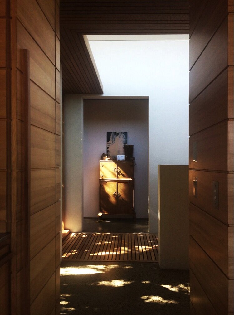 Interior hallway with wooden panel walls, sunlight filtering through the ceiling opening, leading to a small wooden cabinet against the wall, with a piece of artwork above it.