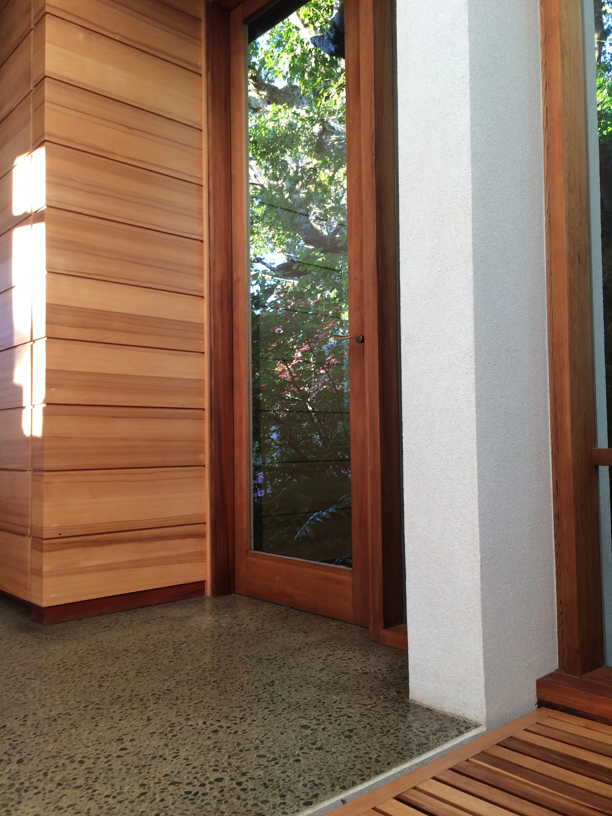 Interior view of a wooden door with glass panel, next to a white textured wall, and a small porch with wooden slats, with green trees visible outside.