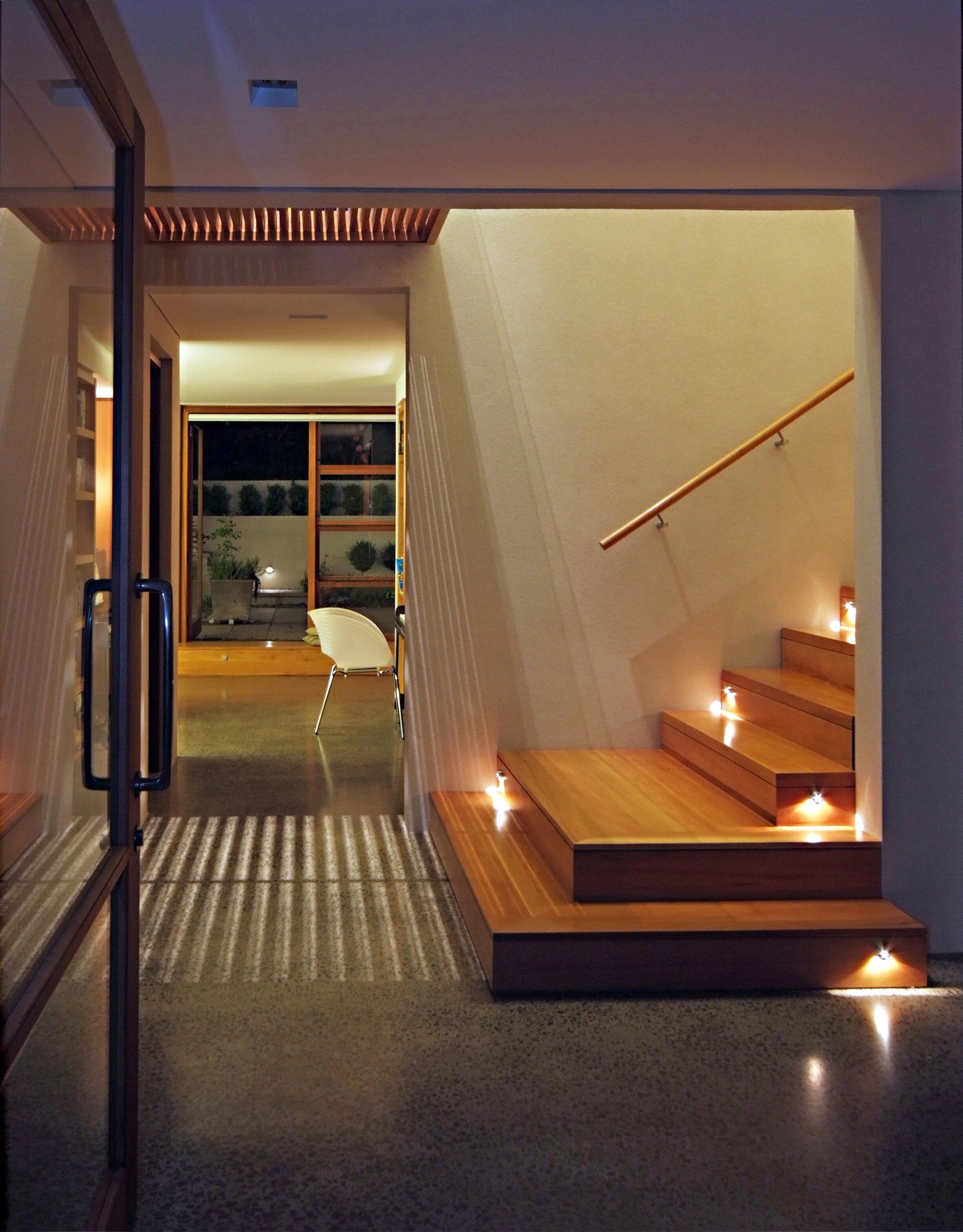 An interior view of a modern house entrance with a wooden staircase lit by small lights, leading to an upper floor, and a sitting area with a chair near large glass doors opening to a patio with potted plants.