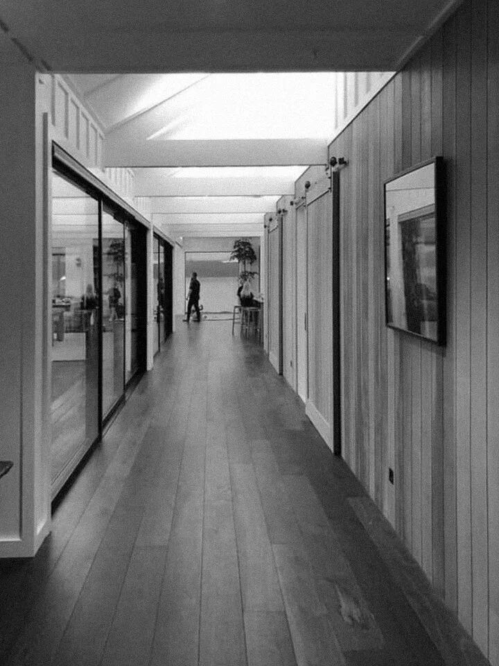 A black and white photo of a corridor with wooden flooring, glass doors on the left, and wood-paneled walls on the right. There are people and plants visible at the end of the corridor.
