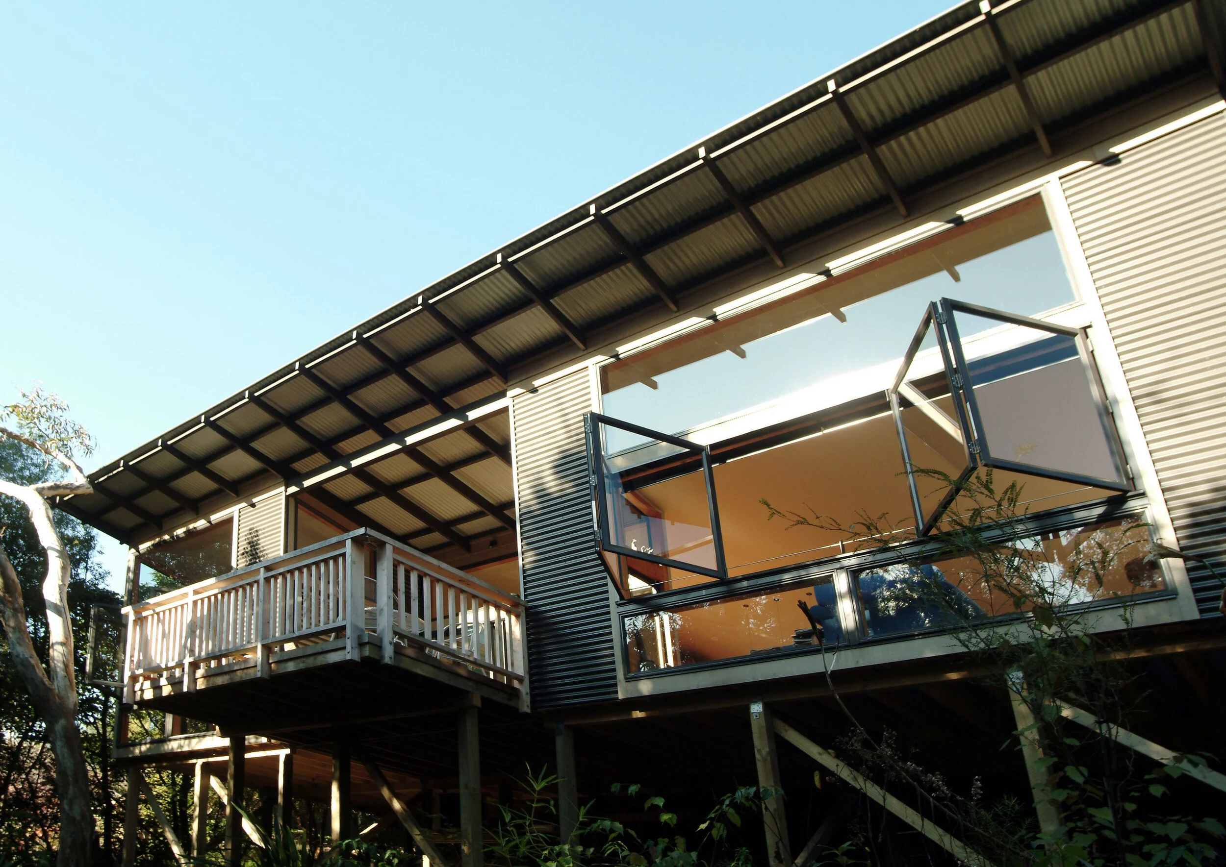 Modern house with large windows and a small balcony, situated among trees with sunlight casting shadows.