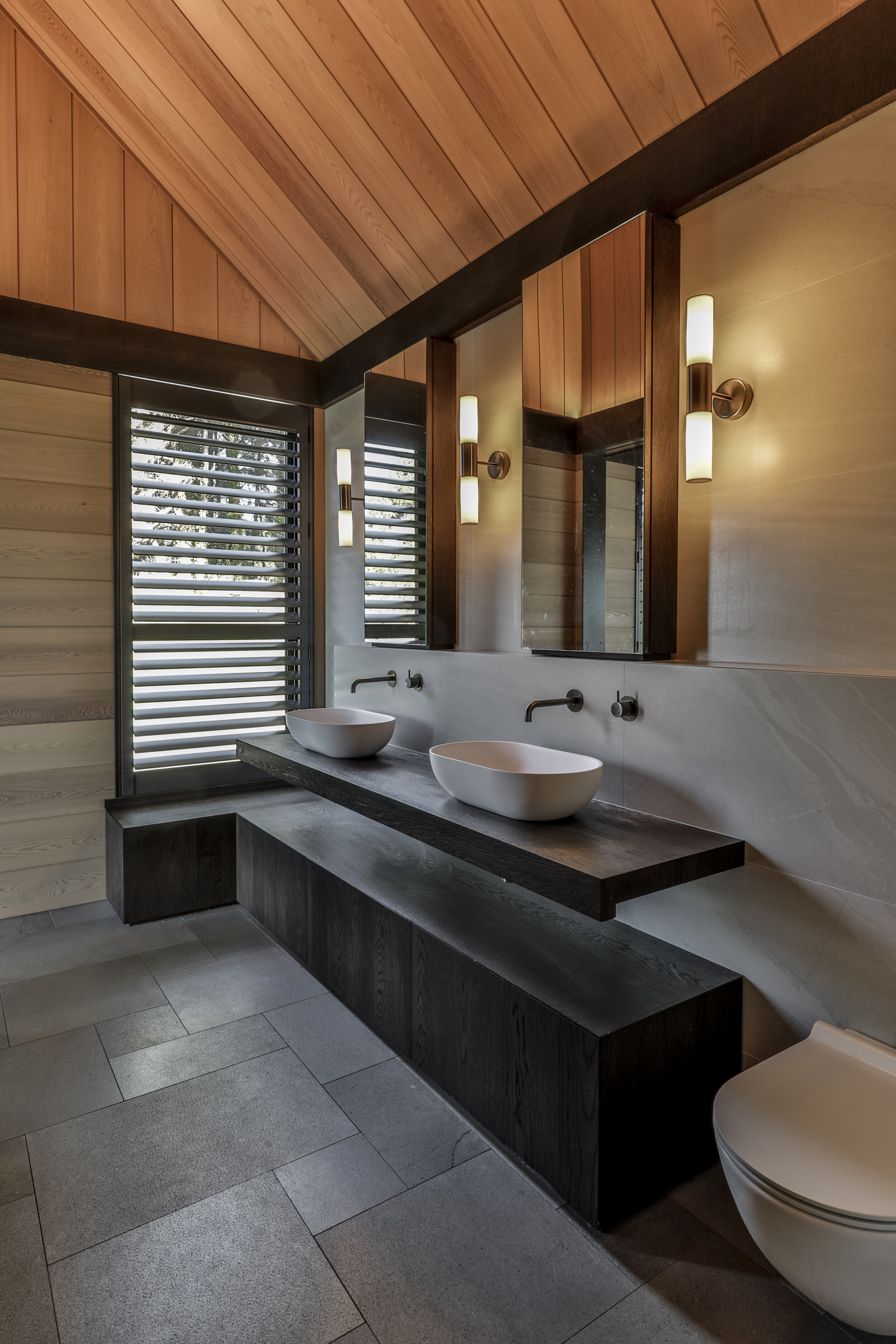 Modern bathroom with wood-paneled ceiling, double vessel sinks on a dark wooden countertop, wall-mounted faucets, and vertical light fixtures, with a window with louvered blinds.