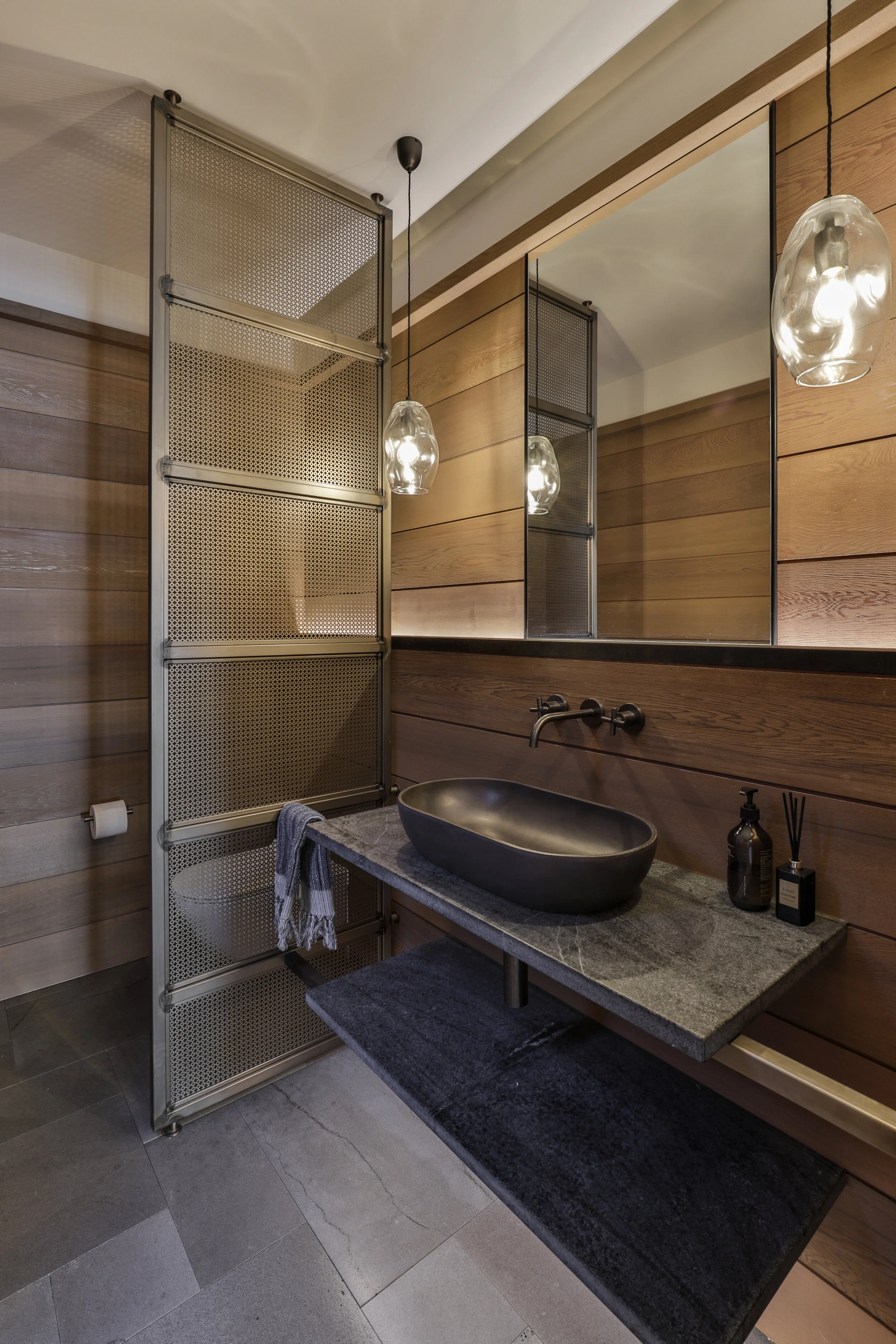 Modern bathroom with wooden panel walls, a large mirror, hanging pendant lights, a stone countertop with a black vessel sink, and a decorative black rug.