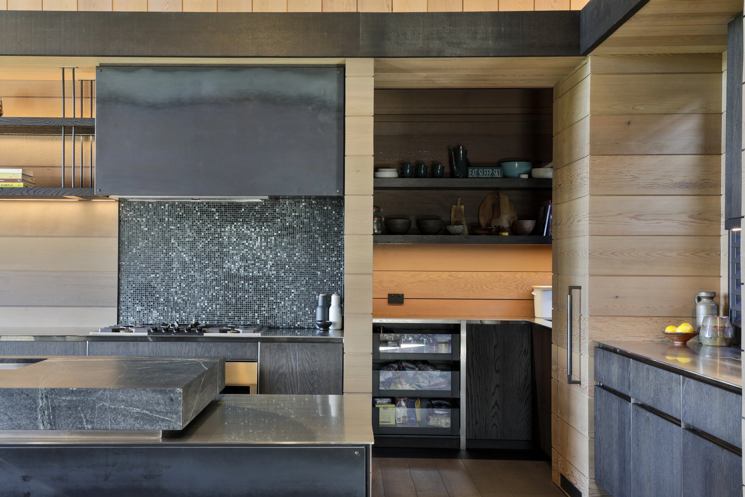 Modern kitchen with wood and dark gray cabinetry, mosaic tile backsplash, open shelves with dishes, and a black countertop with bottles.