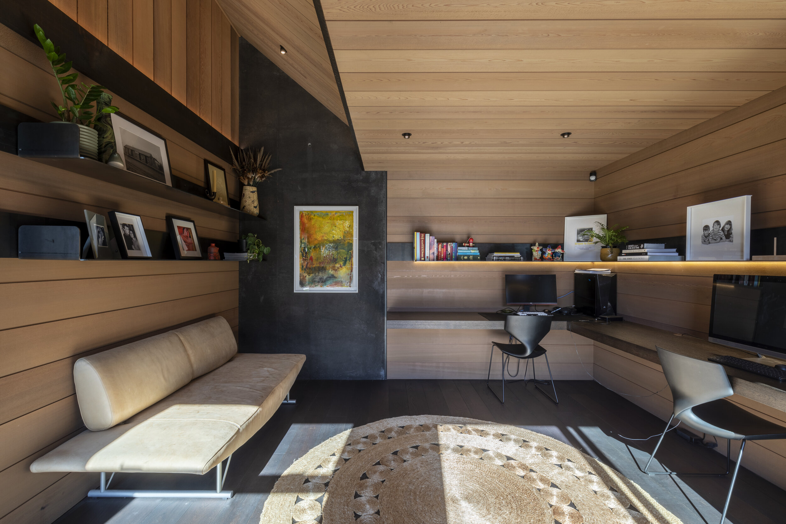 Modern room with wooden walls and ceiling, beige sofa, desk with computer and chair, bookshelves with books, artwork, and framed photographs, rug on dark flooring, plants, and ambient lighting.