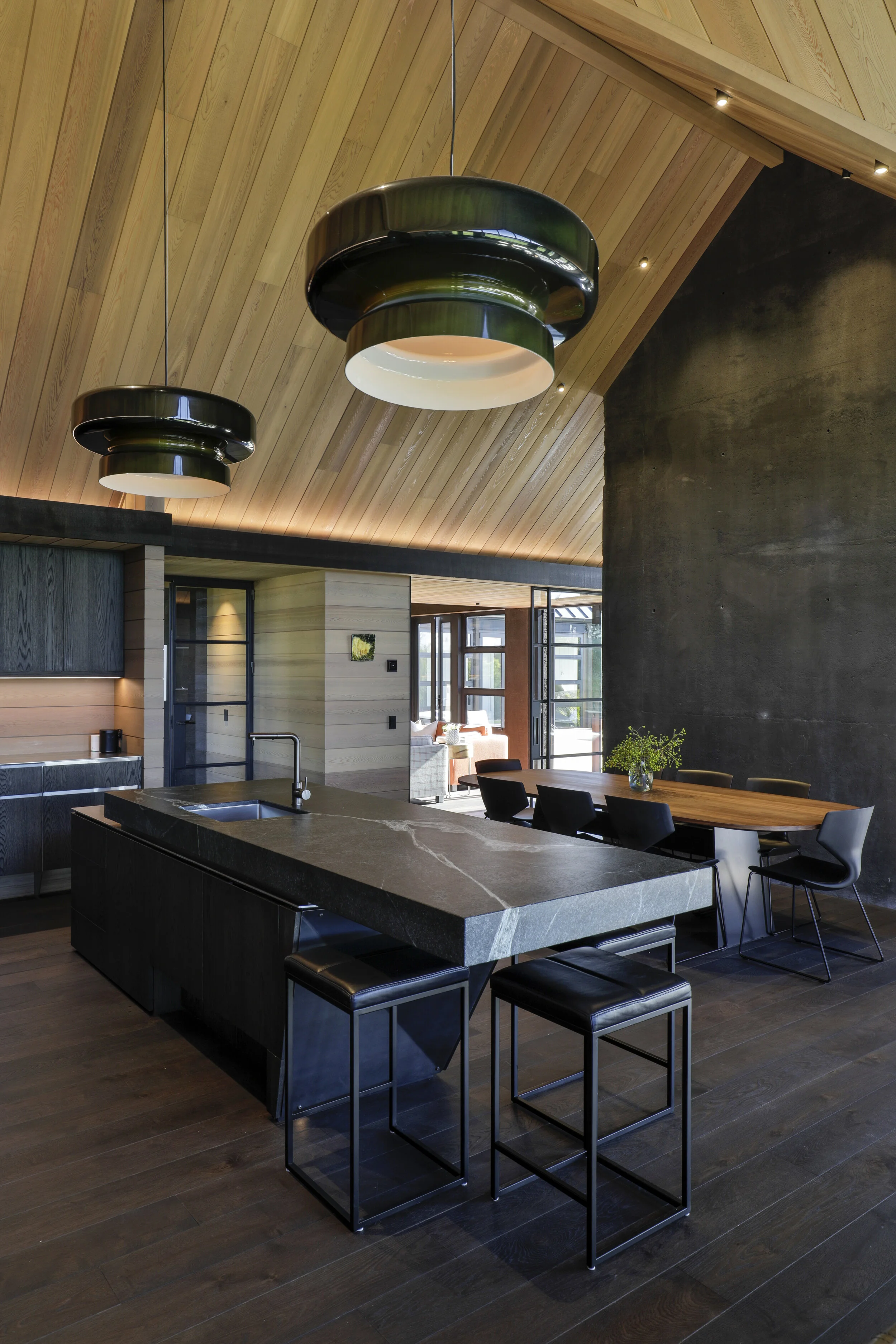 Modern kitchen and dining area with black stone island, wooden ceiling with pendant lights, and large windows.