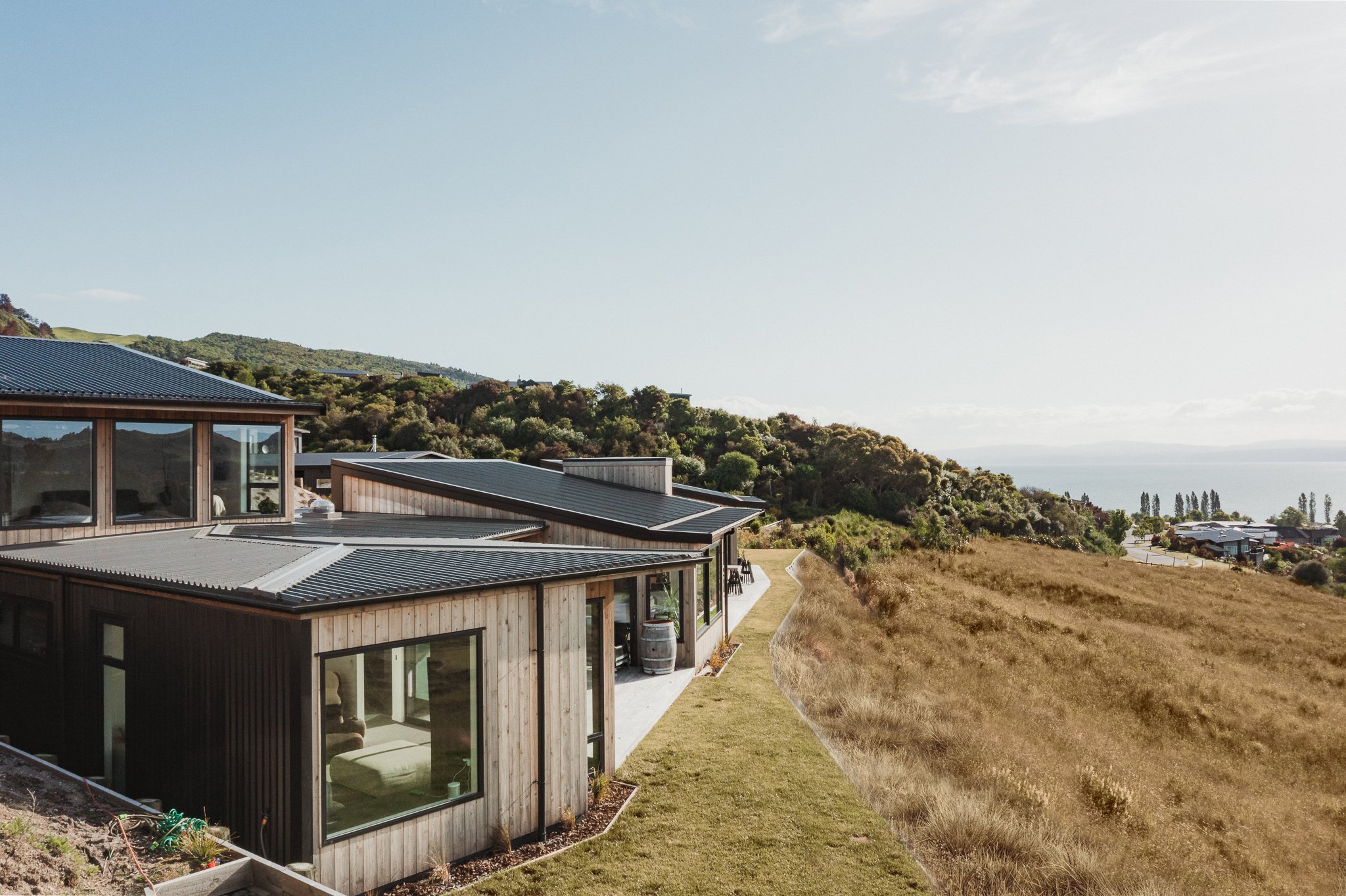 Modern house with wooden and metal exterior, large windows, on a hillside overlooking a grassy field and distant ocean, with a partly cloudy sky.
