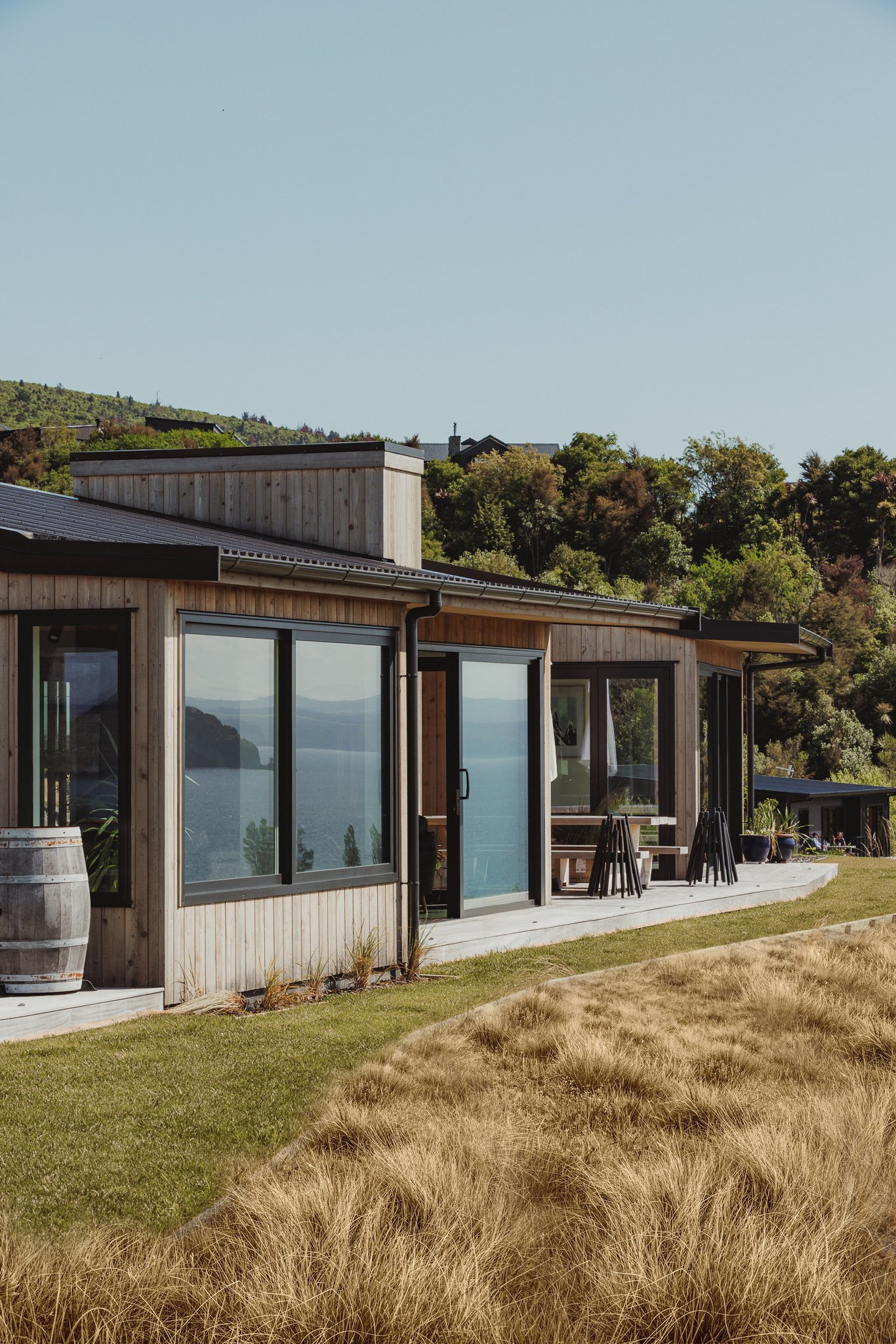 Modern house with large glass windows, wooden exterior, and outdoor patio, overlooking water and hills with trees in the background.
