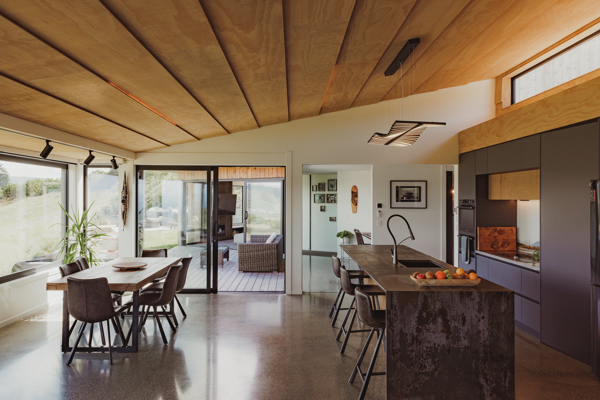Open-concept modern kitchen and dining area with large windows, a wooden ceiling, and outdoor patio seating.