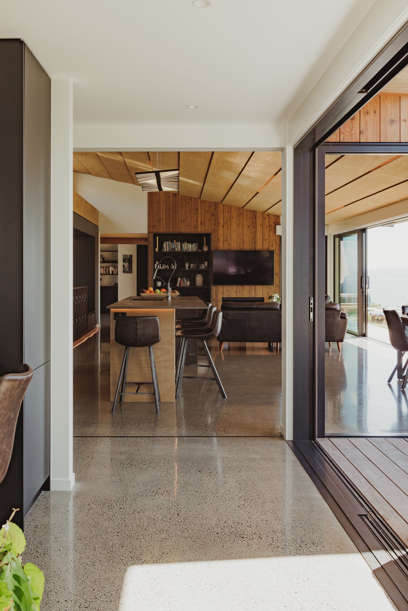Modern open-plan living space with wood-paneled walls and ceiling, a kitchen island with barstools, black bookshelves, a mounted TV, and a sliding glass door leading to an outdoor patio with ocean views.