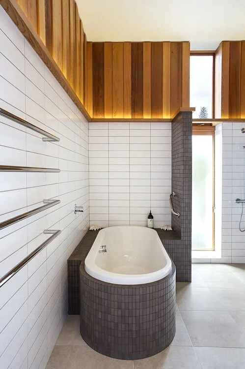 Bathroom with a white bathtub, white tiled walls, wooden ceiling, and glass door with a large window in the background. Designed by Taupo, New Zealand Architects Fraser Cameron Architects. Interior Design by Fraser Cameron Architects. Award Winning B