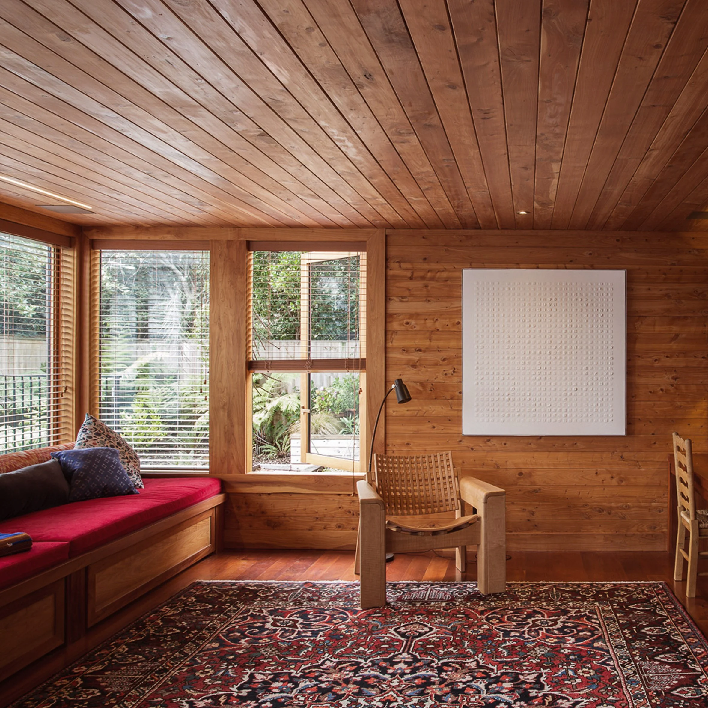 A cozy room with wooden walls, ceiling, and floor. Large windows with blinds allow natural light to fill the space. There is a red-seat built-in window bench with colorful pillows on the left. In the center, a modern wooden chair with a curved design
