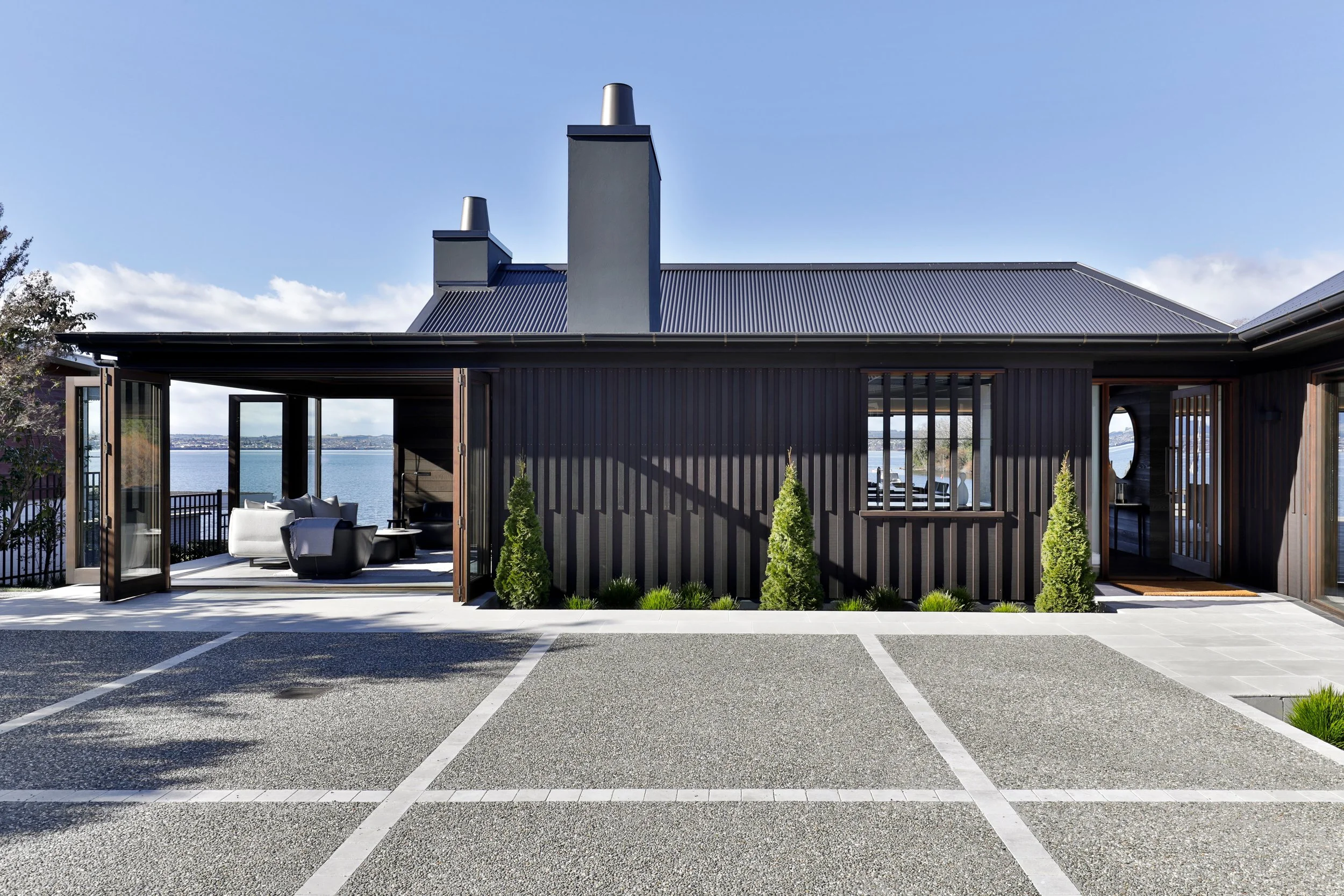 Modern house with black exterior, large open patio area with outdoor seating, overlooking a body of water, with four small shrubs in front. Taupo Architects. The Craftsmen Taupo Builders. Traditional Architects North Island. Traditional Architects So