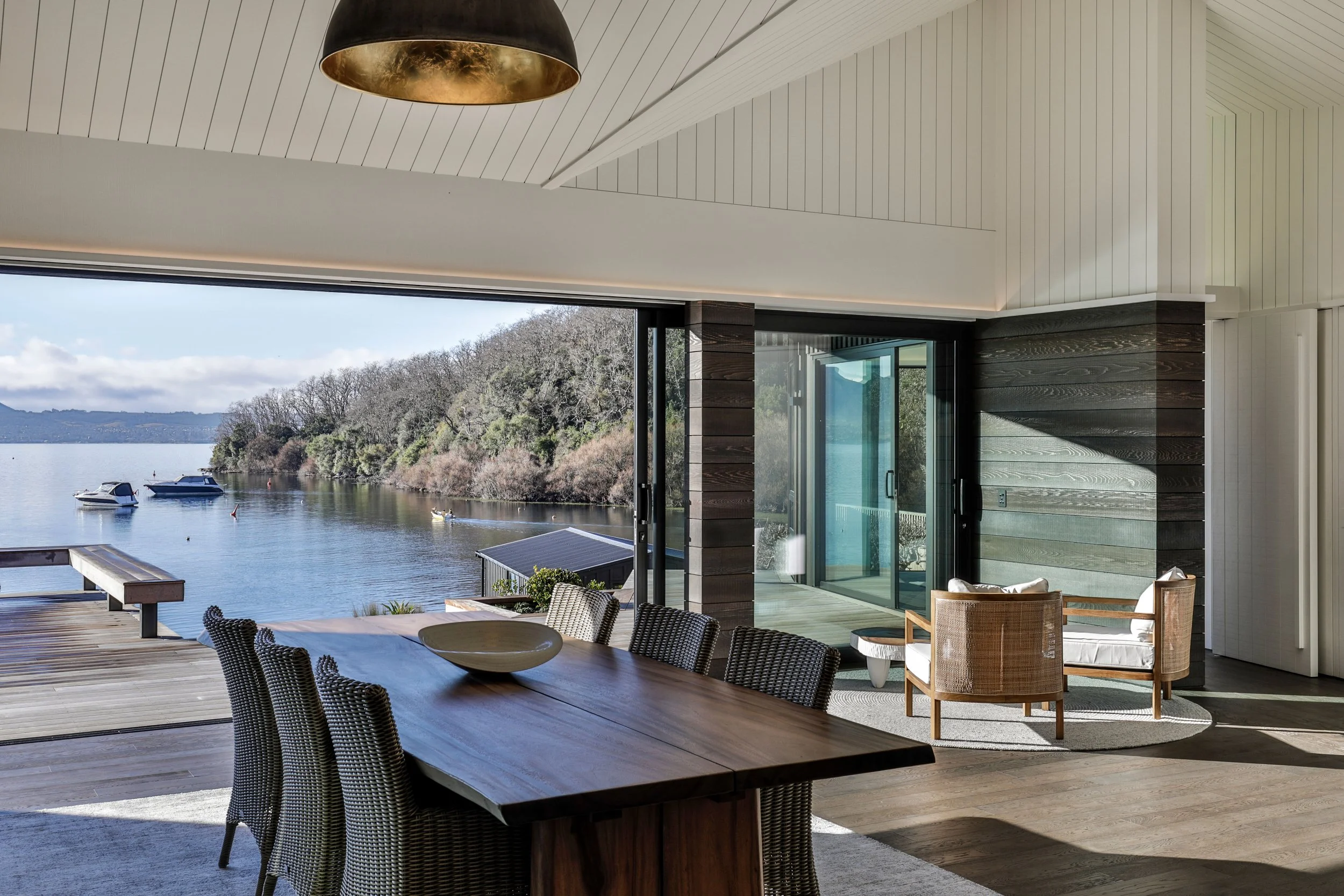 Interior of a modern house with a large open window overlooking a lake with boats and trees, featuring a wooden dining table, wicker chairs, and a sitting area with two wooden chairs and a small table. Taupo Architects. The Craftsmen Taupo Builders. 