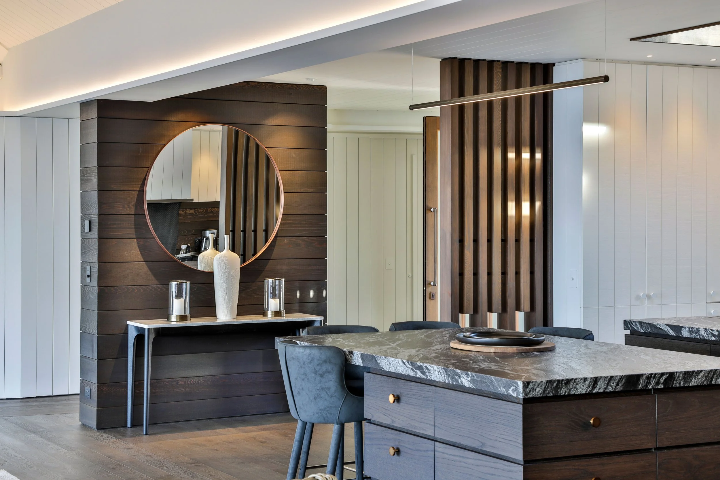 Modern kitchen with a dark wood accent wall, a circular mirror, a marble-topped cabinet with vases and candles, a marble kitchen island, and a dining table with chairs. Taupo Architects. The Craftsmen Taupo Builders. 