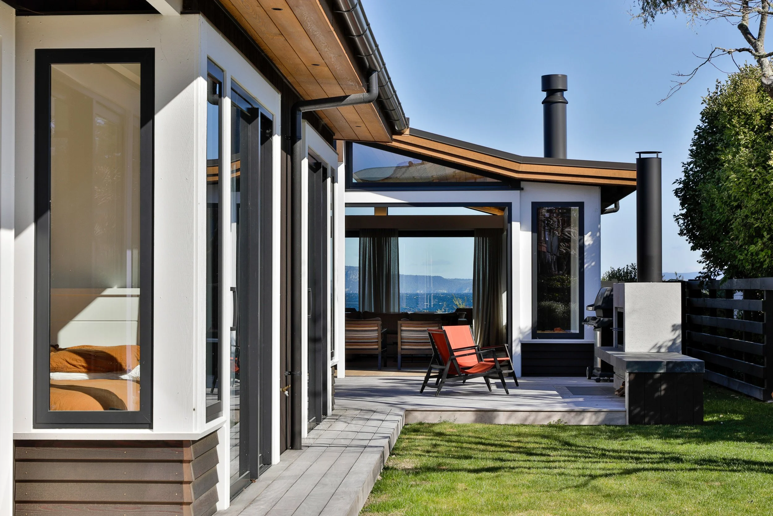 Exterior view of a modern house with large glass windows and a spacious patio. The patio has outdoor furniture, including a red lounge chair and a dining table with chairs. There is a grill on the right side and a well-maintained lawn in the foregrou