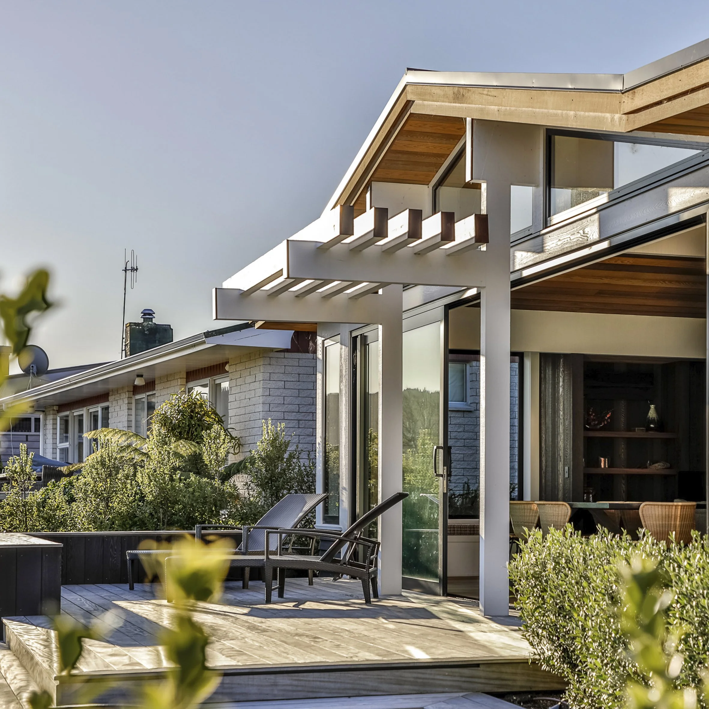 Modern house with a wooden deck, outdoor chairs, and lush greenery, under bright daylight.