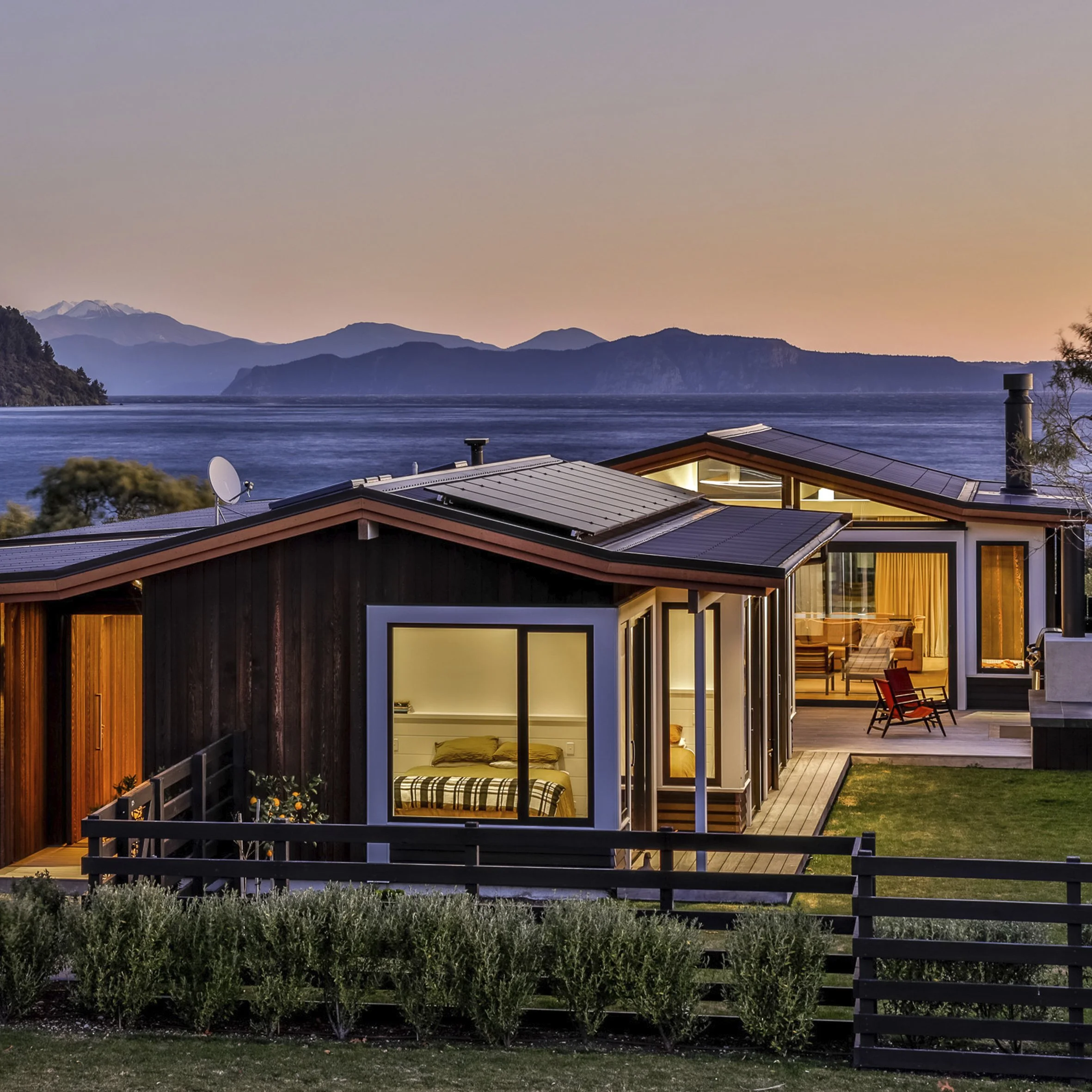 Modern house with large windows overlooking a body of water and mountains at sunset, outdoor patio with furniture.