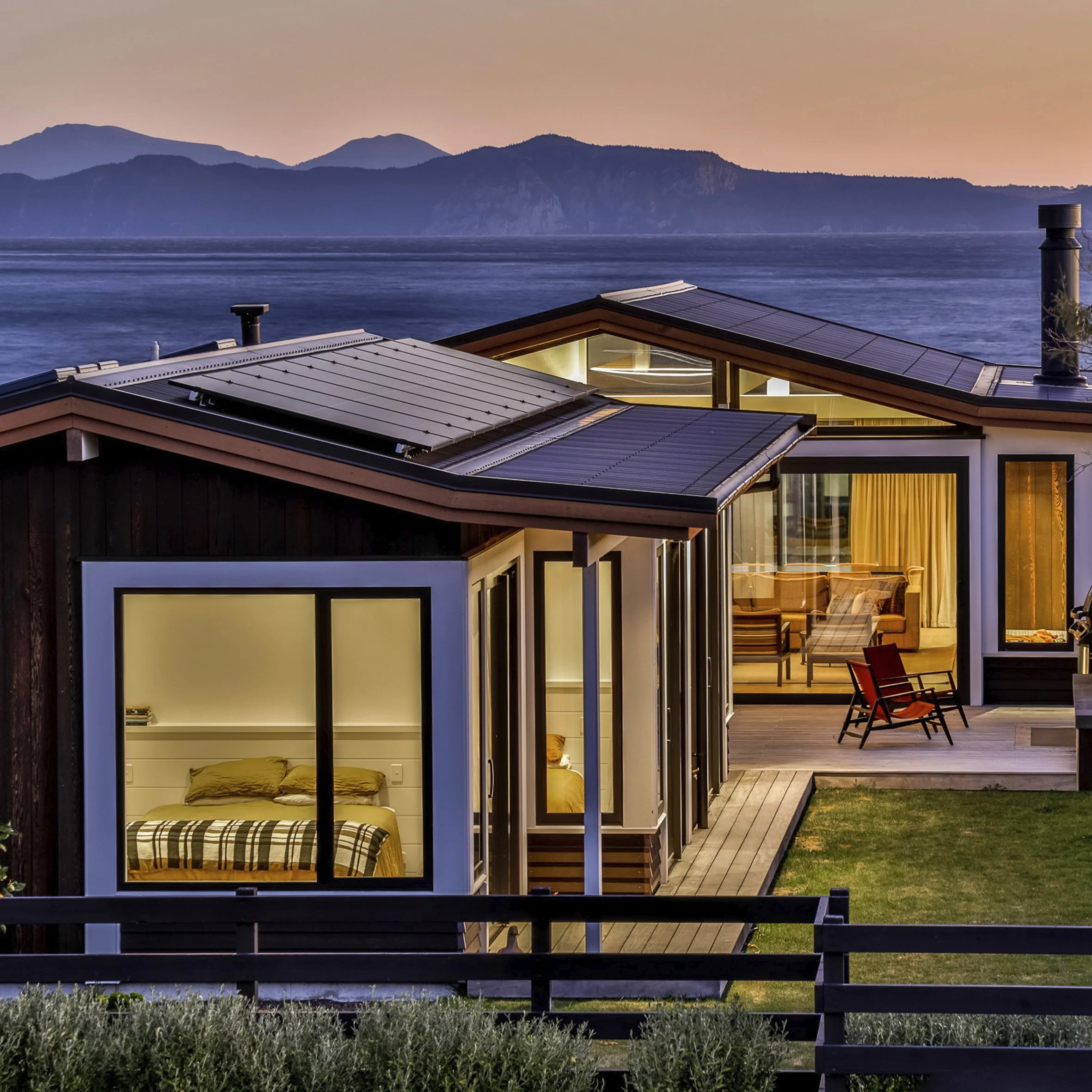 Modern house with large windows overlooking a body of water and mountains at sunset, with interior lighting visible through the windows.