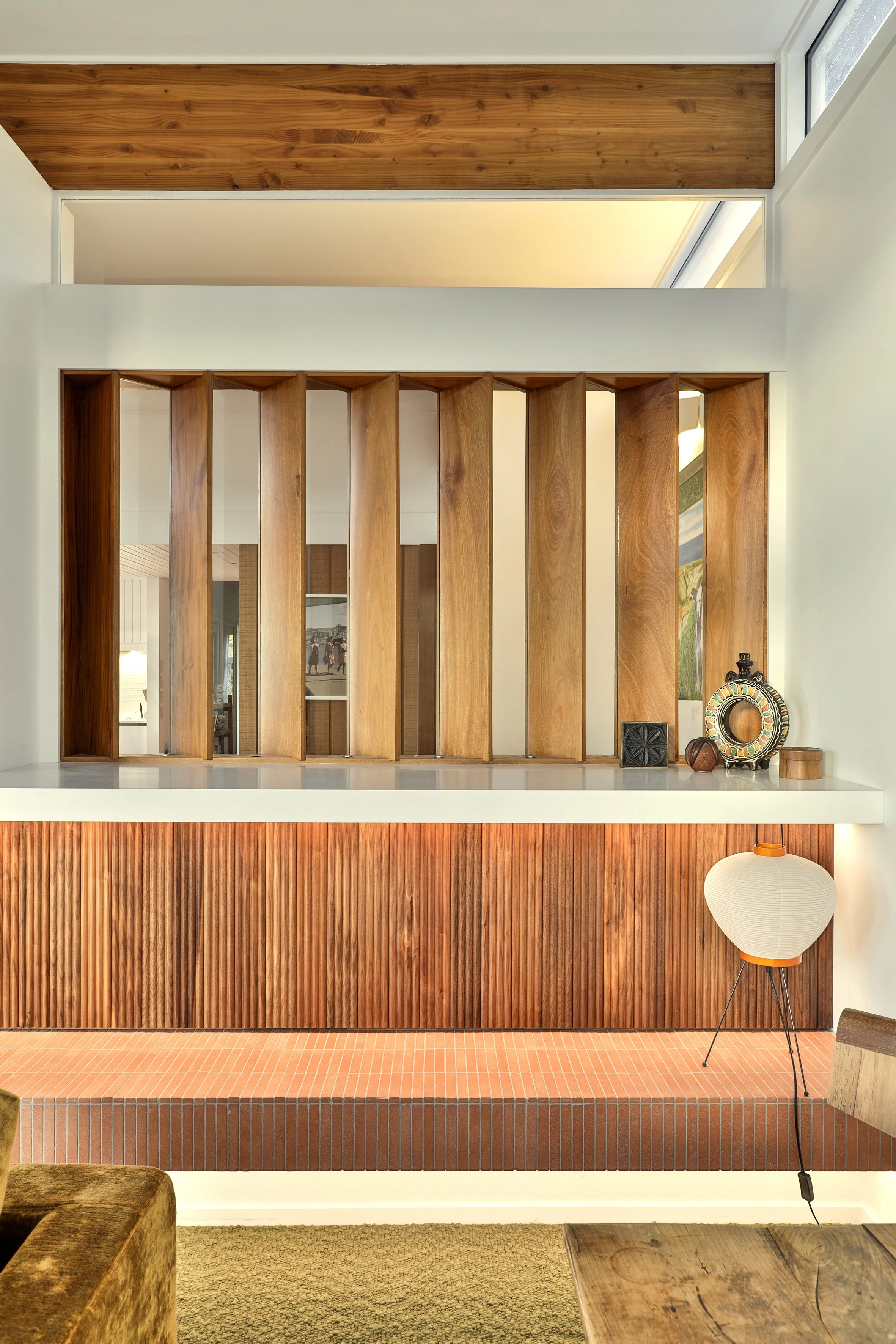 Interior decor featuring a wooden partition with vertical slats, decorative objects on a white ledge, a floor lamp with a paper shade, and a wooden table.