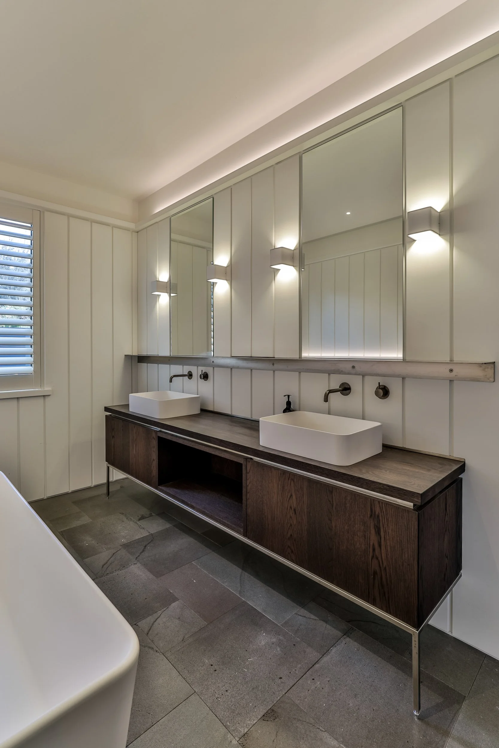Modern bathroom with double white vessel sinks, wide wooden vanity, large mirror, and wall-mounted lights.