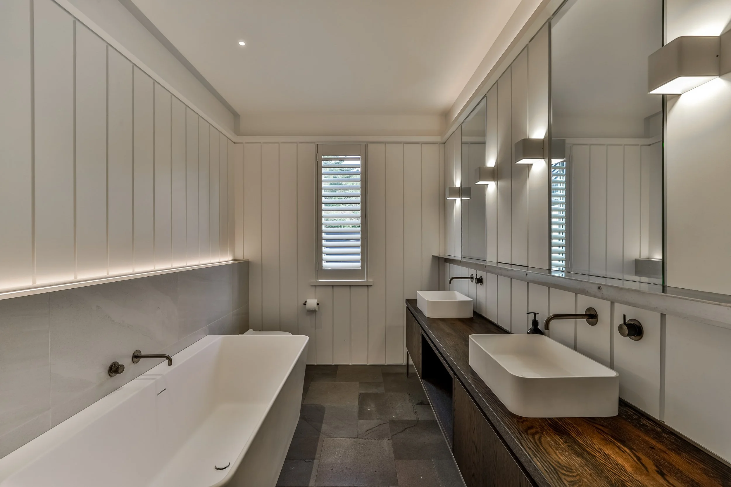 Modern bathroom with a white bathtub, dark wooden vanity with two vessel sinks, large mirror, and a small window with shutters.