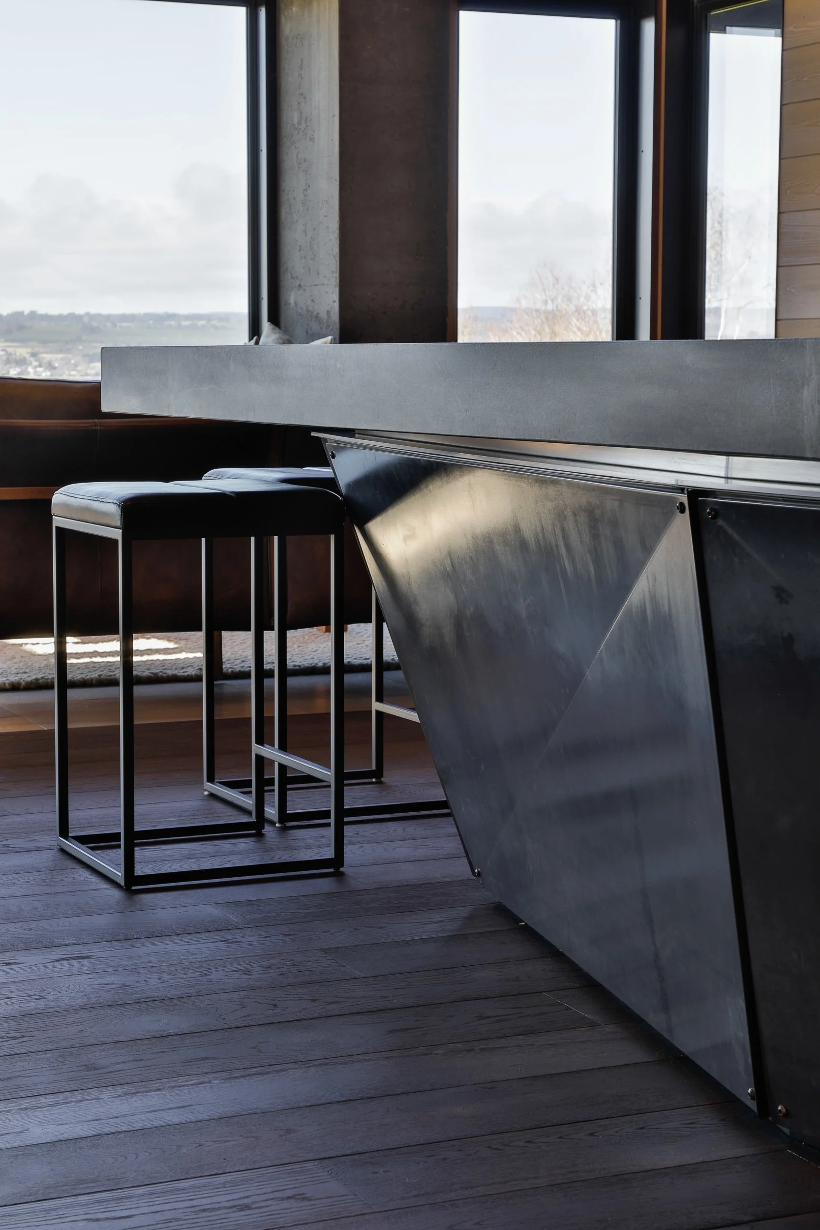 Modern interior with a large window, black barstools, a concrete countertop, and a sleek metal base on the kitchen island, with wooden floors.