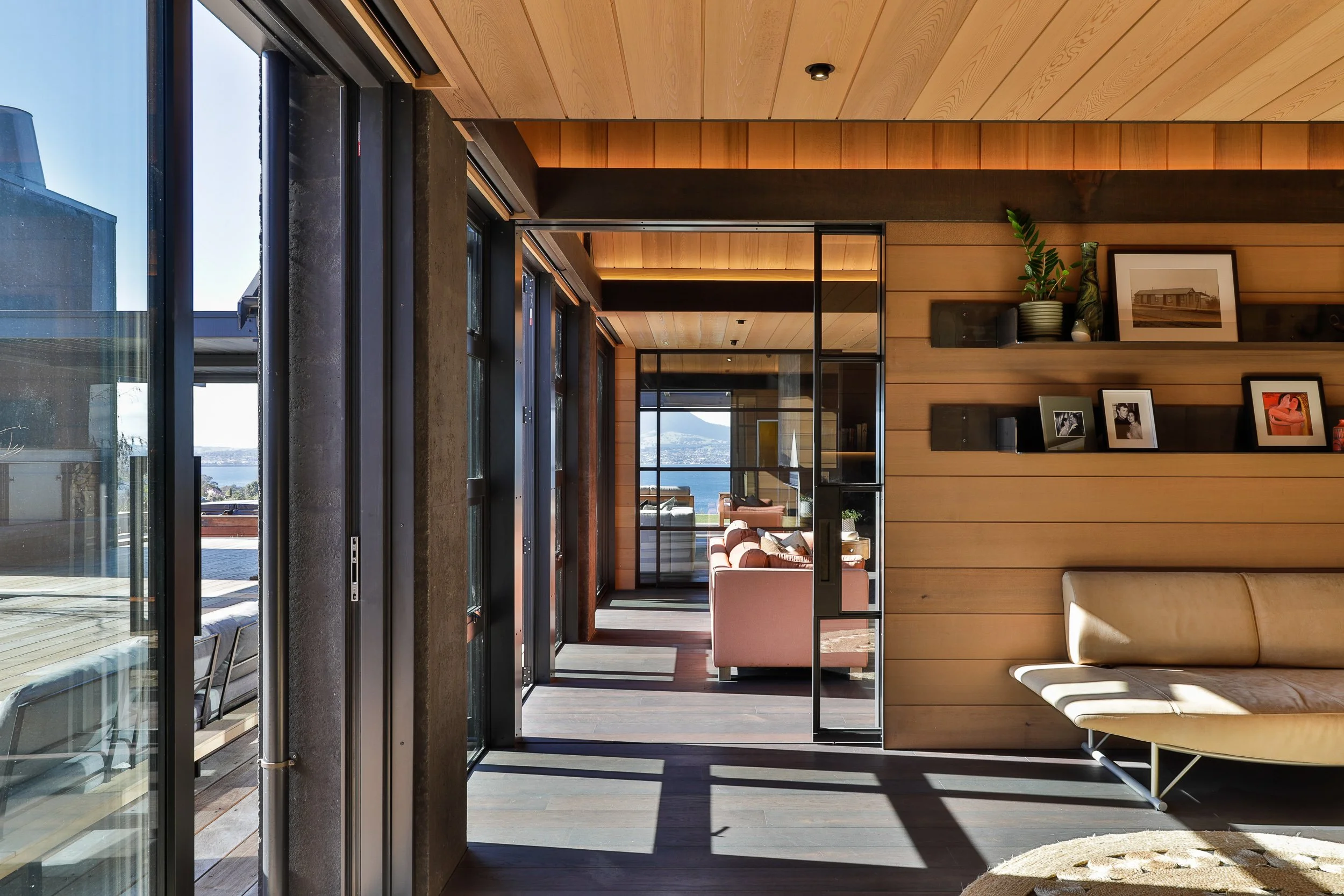 A modern living room with wooden panel walls, glass sliding doors leading outside, and a spacious interior with a beige couch and wall-mounted shelves with framed photos and plants. Sunlight streams through the glass doors, casting shadows on the dar