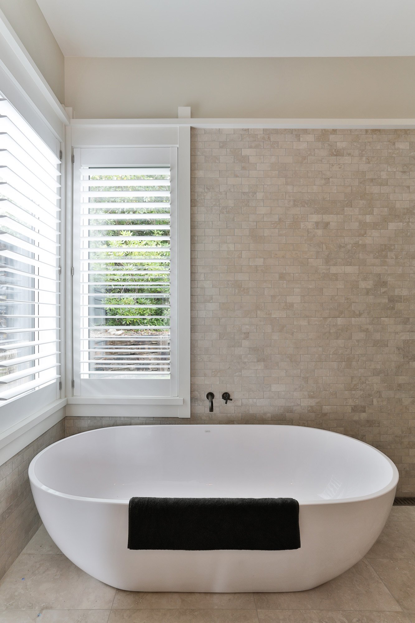 Modern white bathtub with a black towel hanging over its side, positioned in a bathroom with beige tile walls and large windows with white shutters.