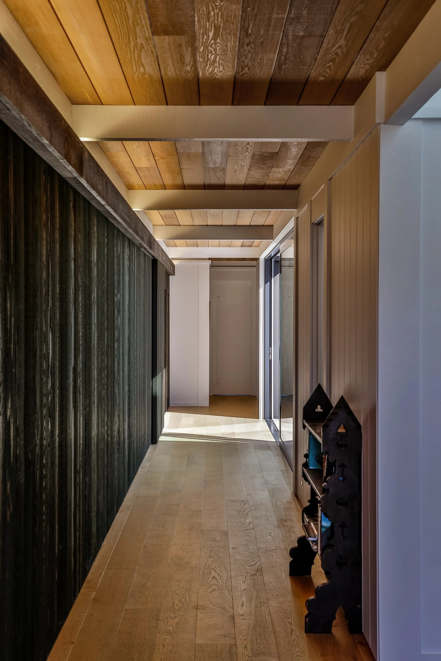 A hallway with wooden flooring and a wood-paneled ceiling, featuring a black decorative piece on the right side and a sliding glass door at the end.