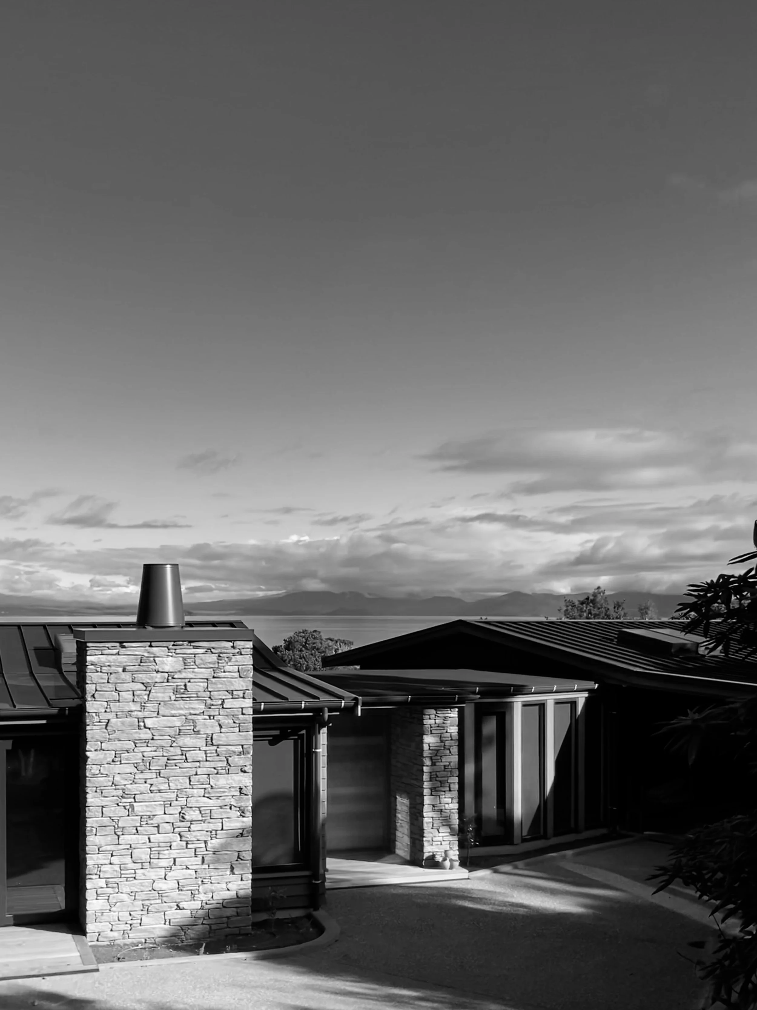 A black and white photo of a modern house with stone and glass walls, metal roofing, and a chimney, with mountains and a cloudy sky in the background.
