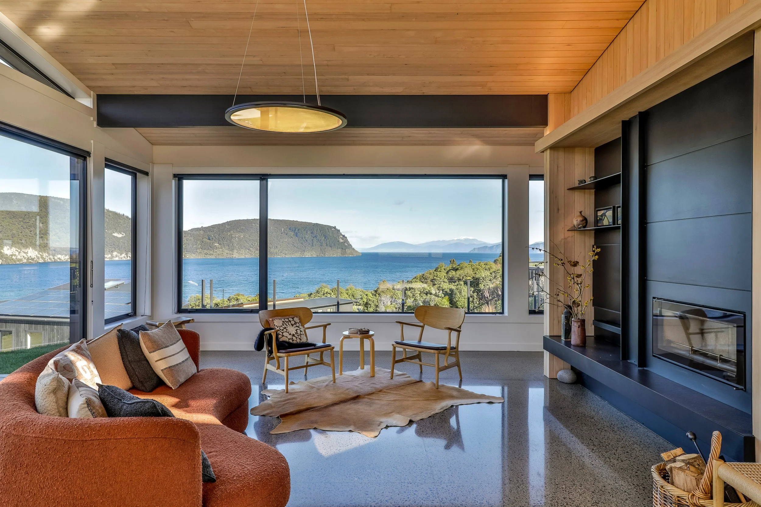 Living room with large windows overlooking a lake and mountains, furnished with a red sofa, wooden chairs, and a black fireplace, with a cowhide rug on a polished concrete floor.