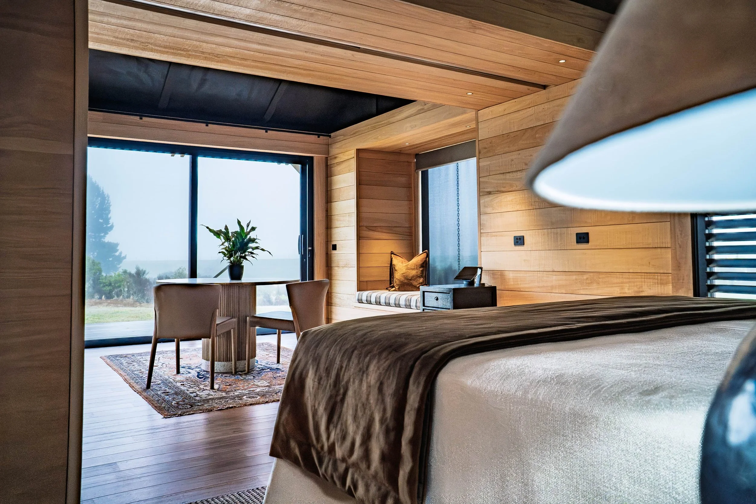 A cozy bedroom with wooden walls and floors, featuring a large bed covered with a blanket, a round table with two chairs near a large glass sliding door, and a window seat with a pillow, overlooking a foggy outdoor landscape.