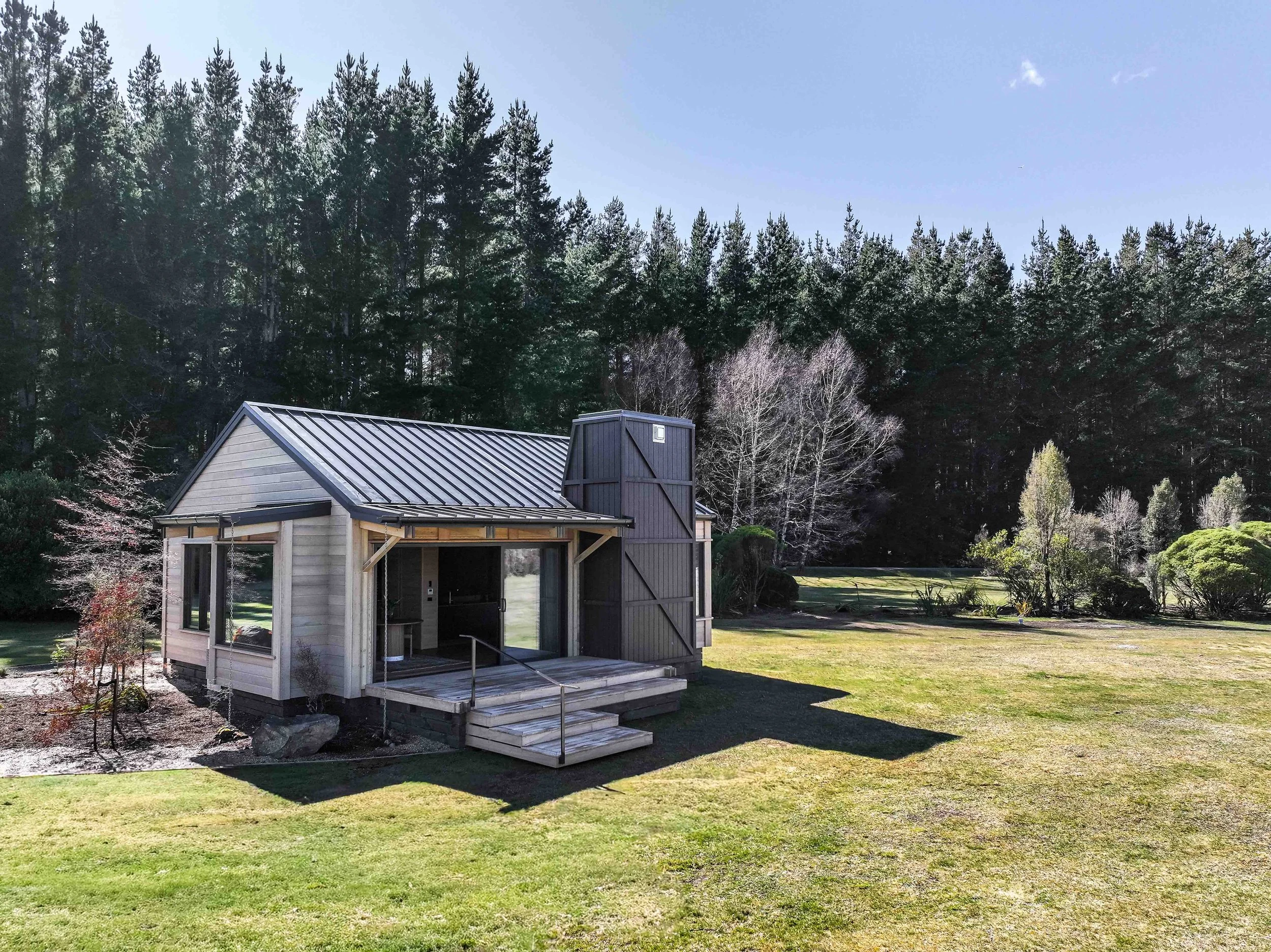 Small wooden house with a metal roof, set in a grassy yard with trees and forest in the background. Lodge architect. Commercial accomodation designer. Hotel designer. Luxury lodge design. Poronui. 