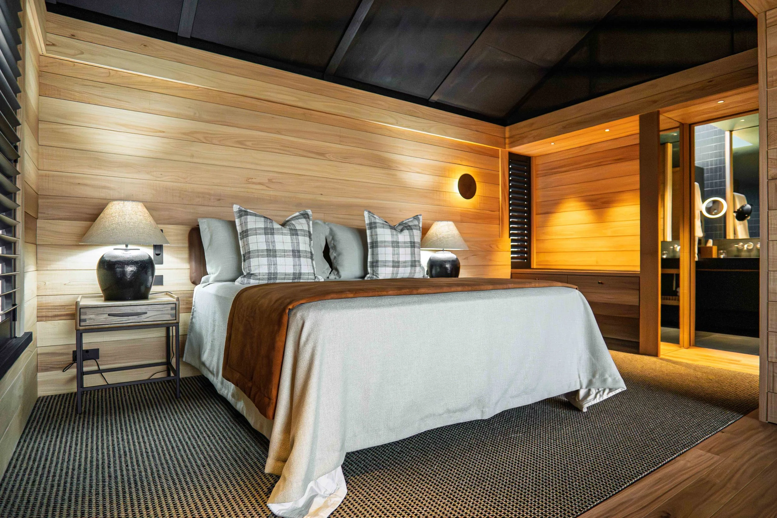 A bedroom with wooden panel walls, a bed with white and plaid pillows, a brown blanket, two black bedside lamps, a window with black blinds, and a doorway leading to a bathroom with black tiles and modern fixtures. Lodge architect. Commercial accomod