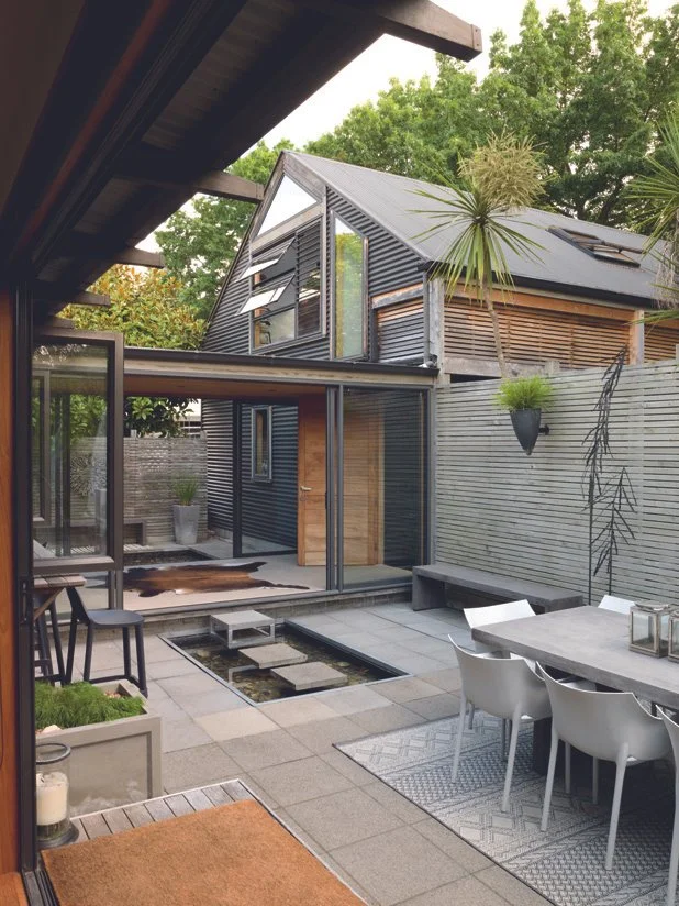 Modern backyard with outdoor seating, small pool, potted plants, and a black architectural style house with large windows and a sloped roof. Decor Design Taupo. Trace Interior Designer Taupo. 