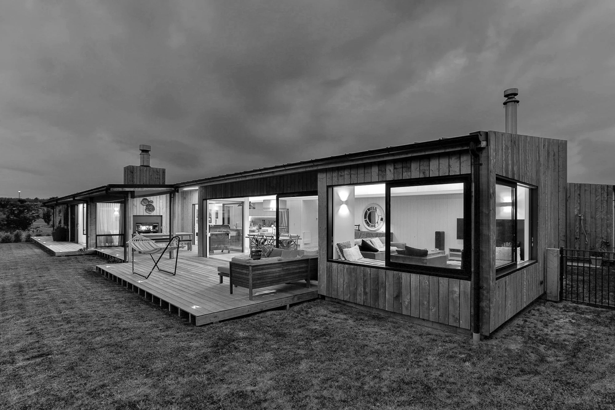 A modern wooden house with large glass windows and a wooden deck, lit from inside, under a cloudy sky.