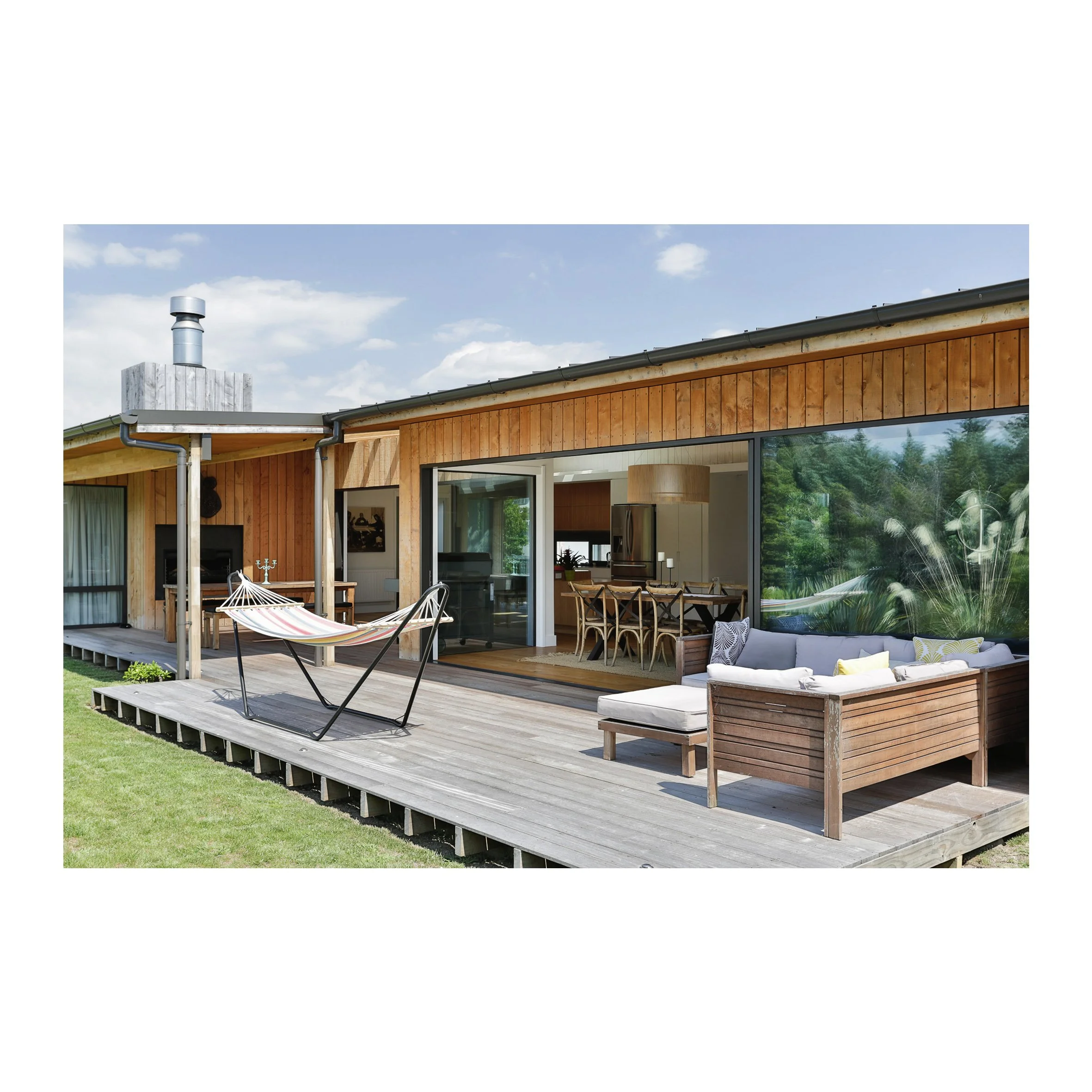 Outdoor view of a modern wooden house with a spacious deck, hammock, outdoor sofa, and dining area indoors visible through sliding glass doors.