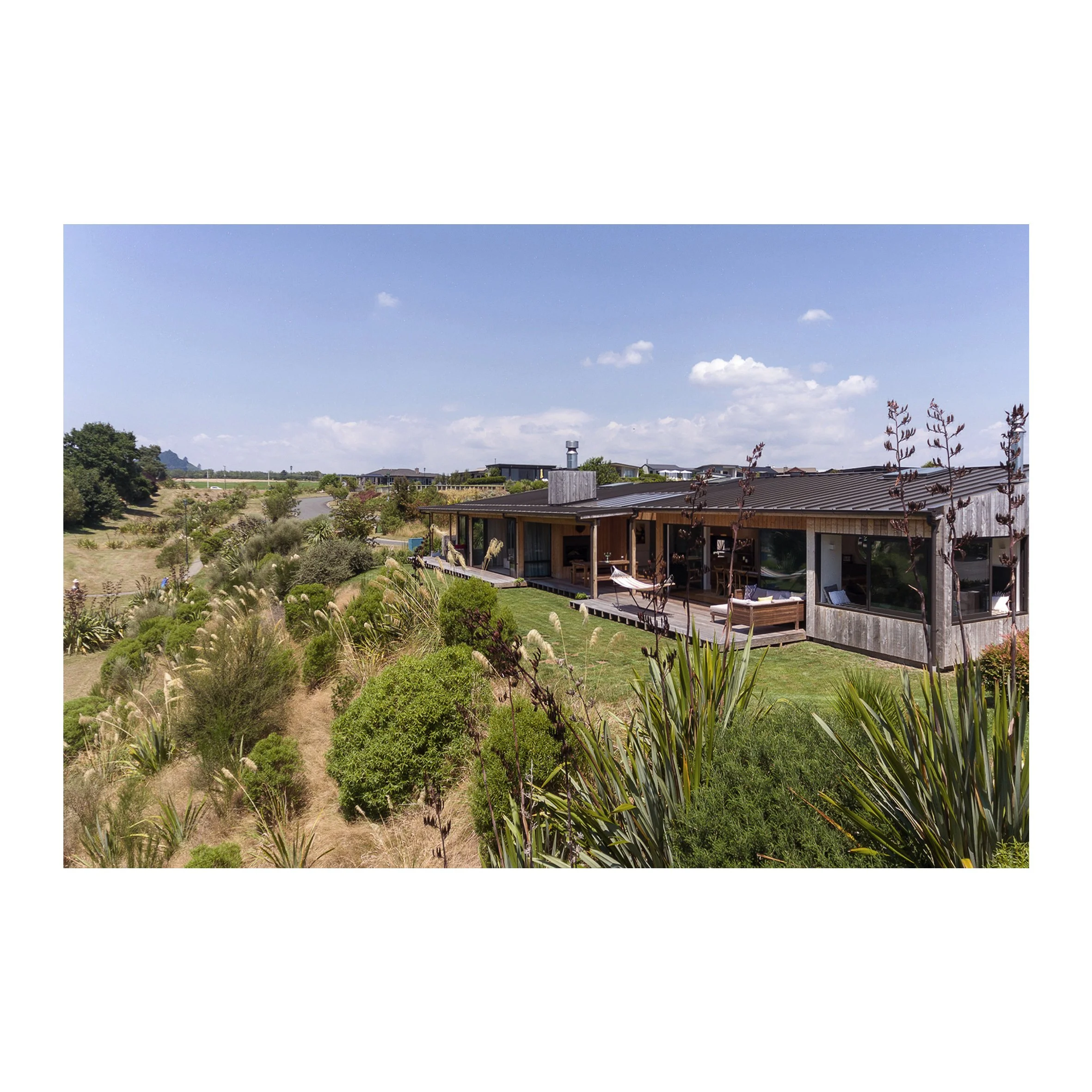 Modern house with a wooden deck on a landscaped hill, surrounded by greenery and plants, under a partly cloudy sky.
