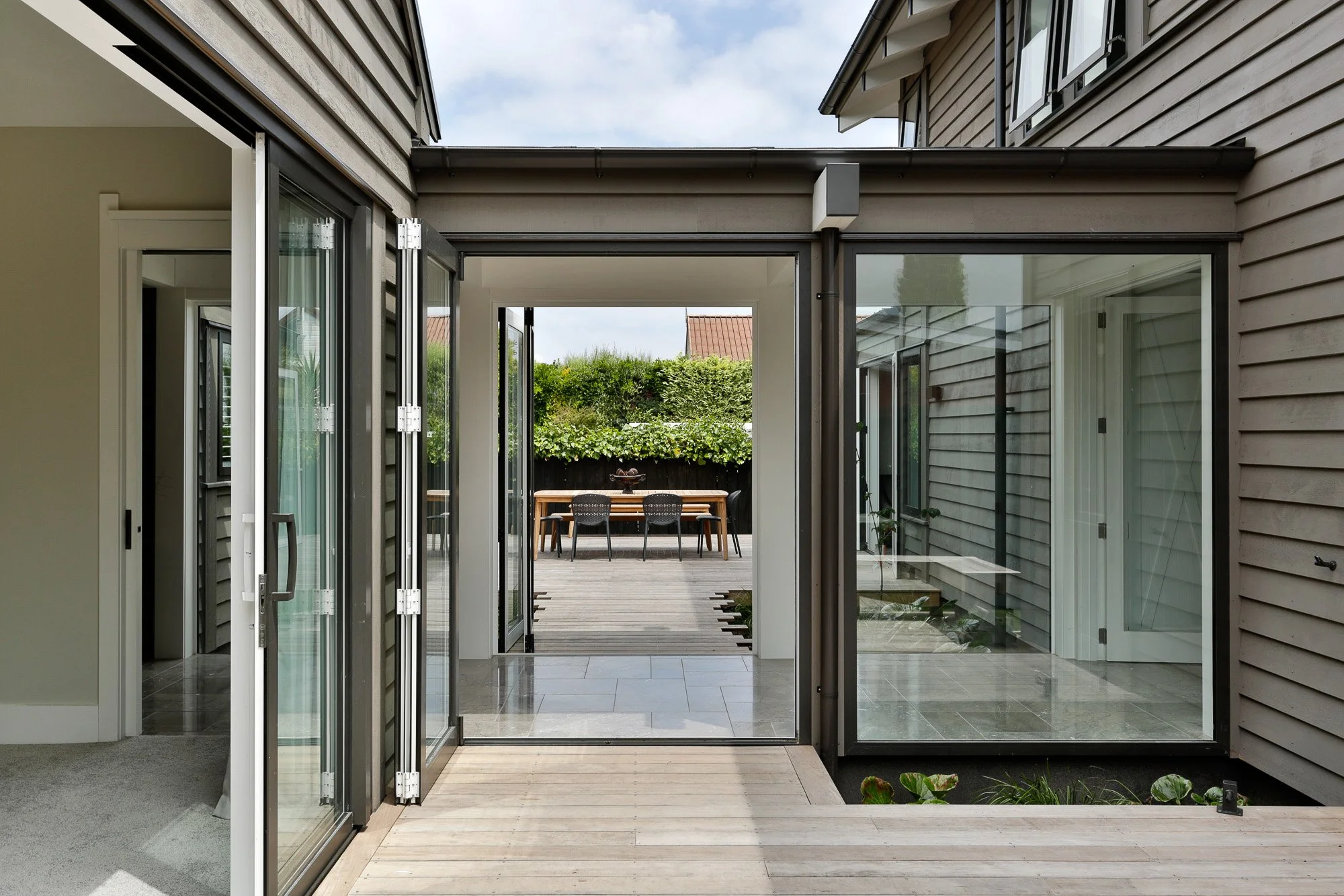 View of a sunlit outdoor terrace with a wooden table and chairs, seen through large glass doors from inside a modern house.