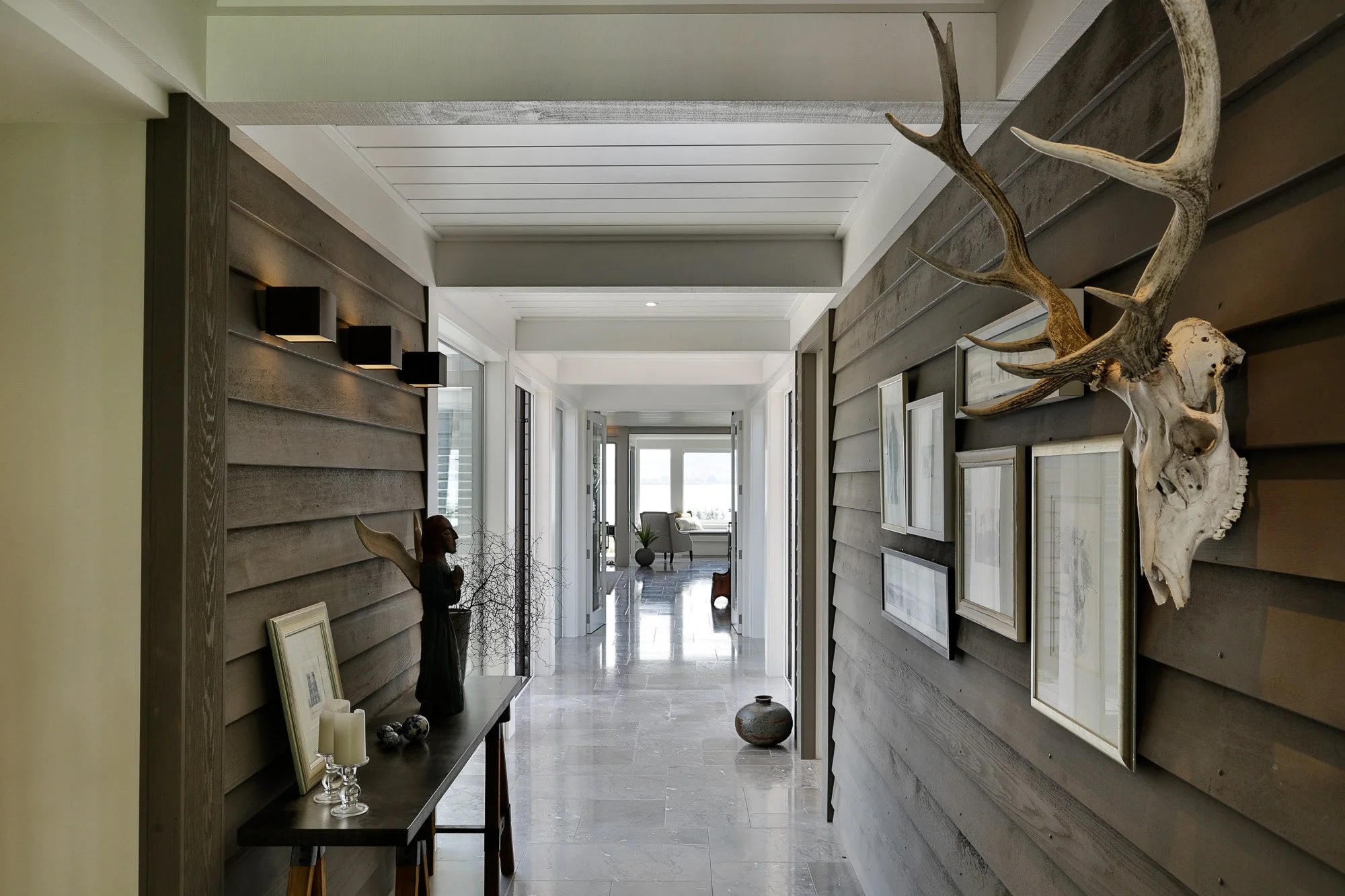 Modern hallway with gray wooden wall panels, art frames, and a mounted deer skull with antlers on the right wall. On the left, a black console table holds candles, a sculpture, and decorative objects. The hallway leads to a bright room with large win