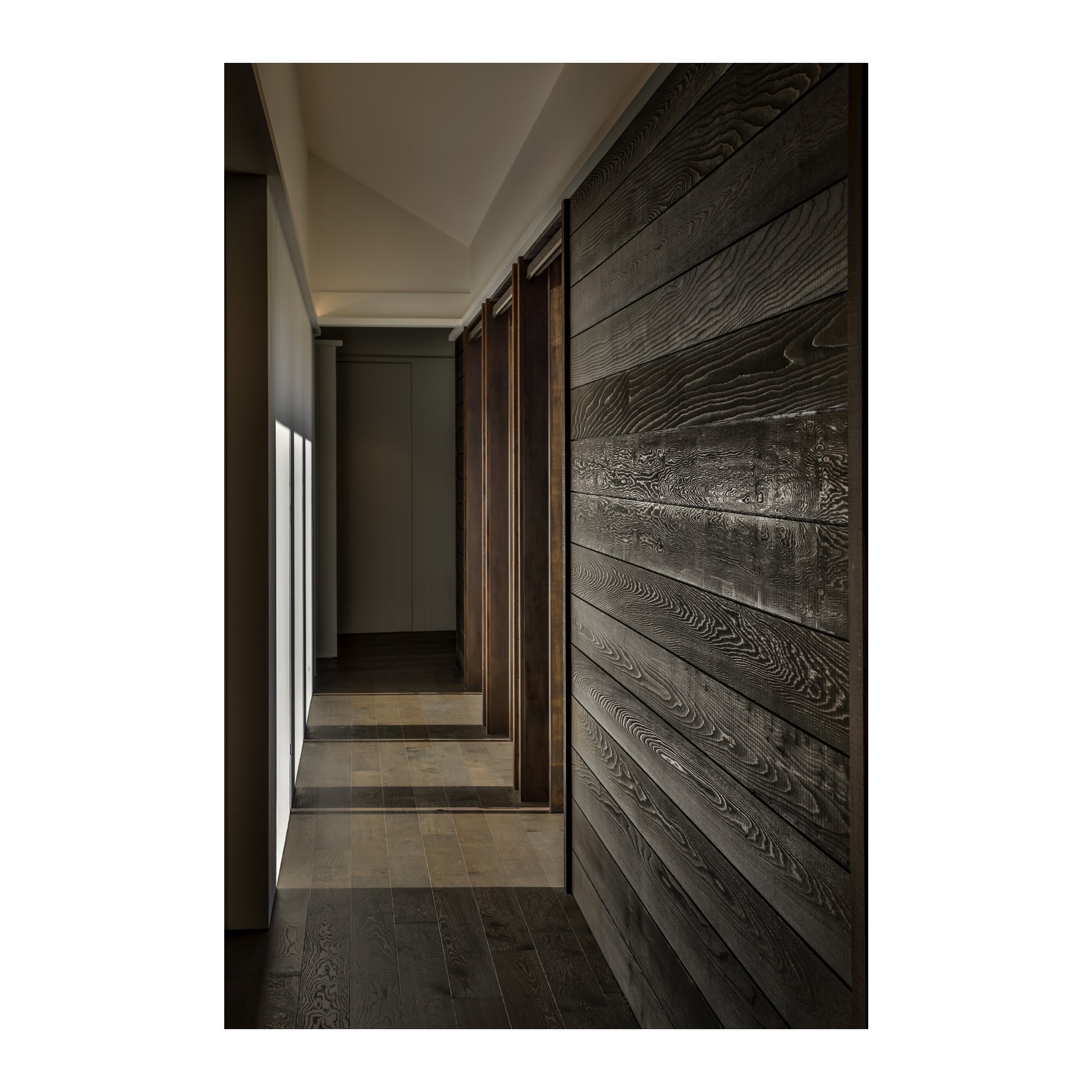 Interior hallway with dark wood paneling on the wall, light switch and shadow pattern on the floor, and natural light coming through a window.