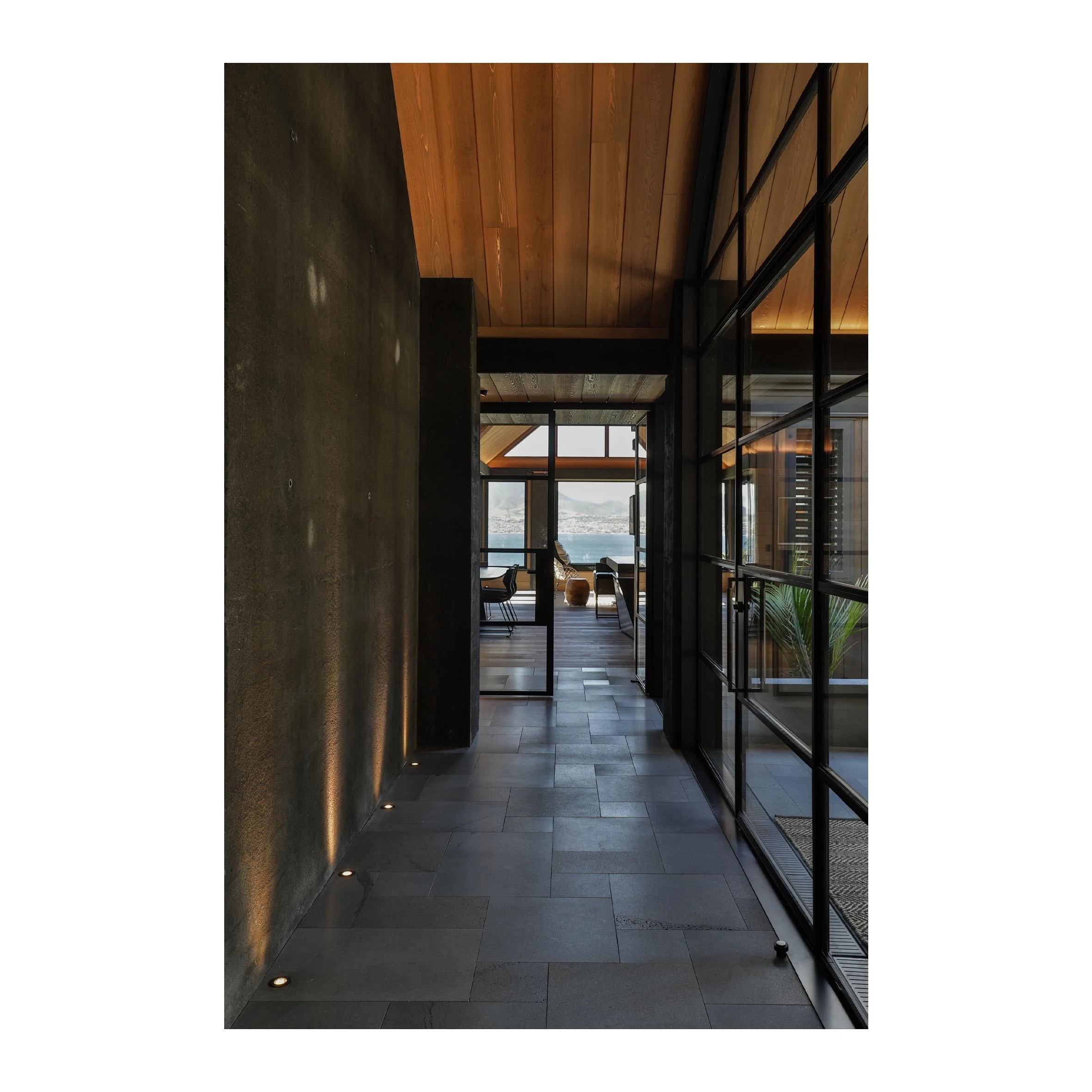 Modern interior hallway with brick and glass walls, wooden ceiling, and view of outdoor landscape through glass door at the end.
