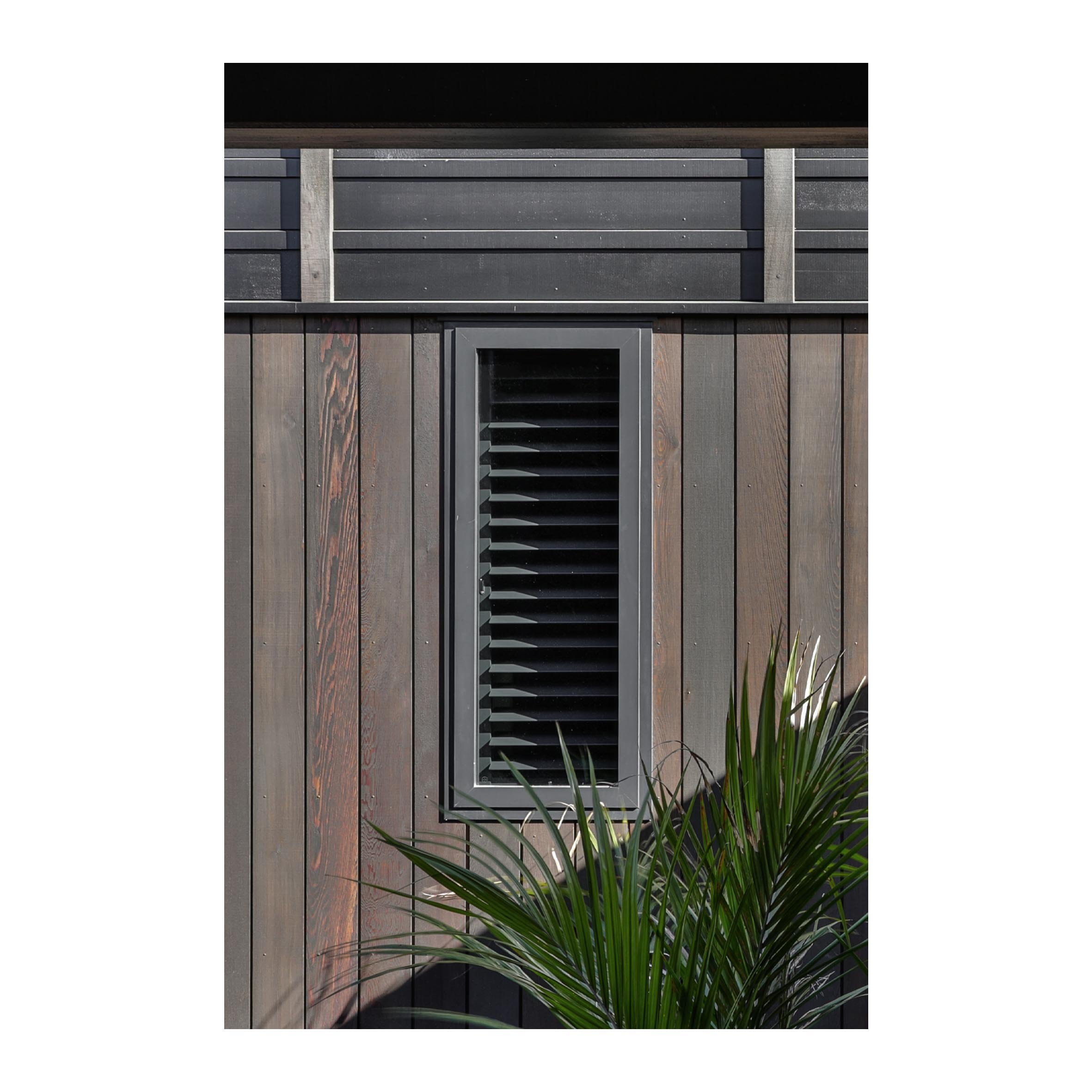 A rectangular black vent on a wooden wall with horizontal slats, with green plants in the foreground.