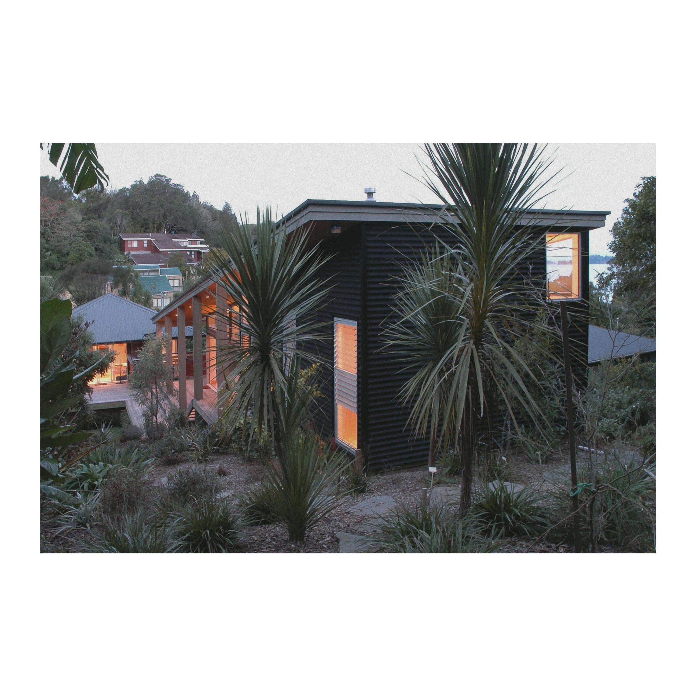 Modern black house with large windows and warm interior lighting, surrounded by desert plants and trees, on a hillside at dusk