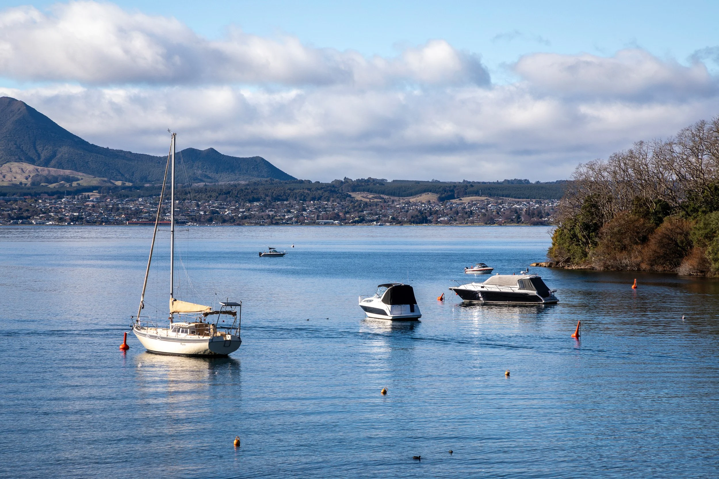 Calm water with several boats anchored, trees on the right, mountain range in the background, partly cloudy sky. Taupo Architects. The Craftsmen Taupo Builders. 